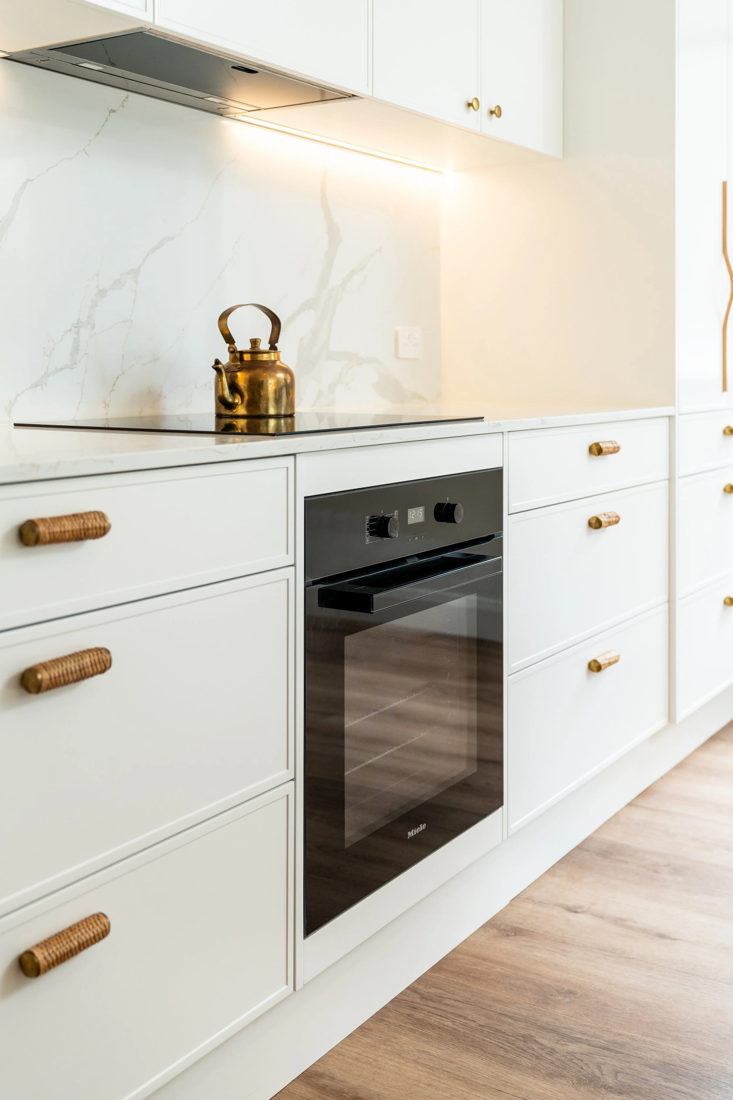 White kitchen with marble backsplash, brass hardware on cabinets, black oven, and brass kettle on stovetop, wooden floor.