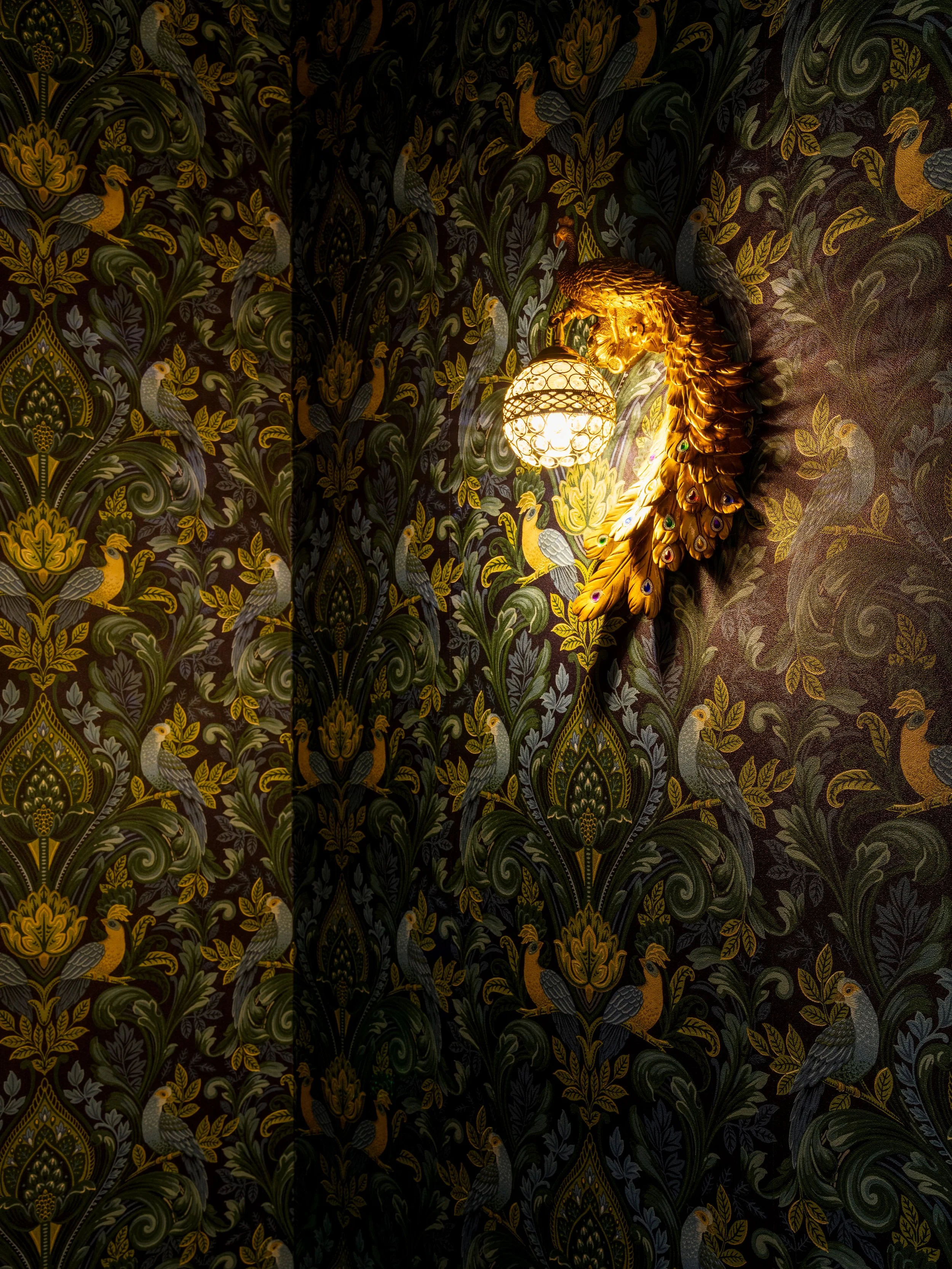 An ornate wall sconce in the shape of a peacock, mounted on wallpaper with a repeating pattern of peacocks and floral motifs, illuminated with a warm light.