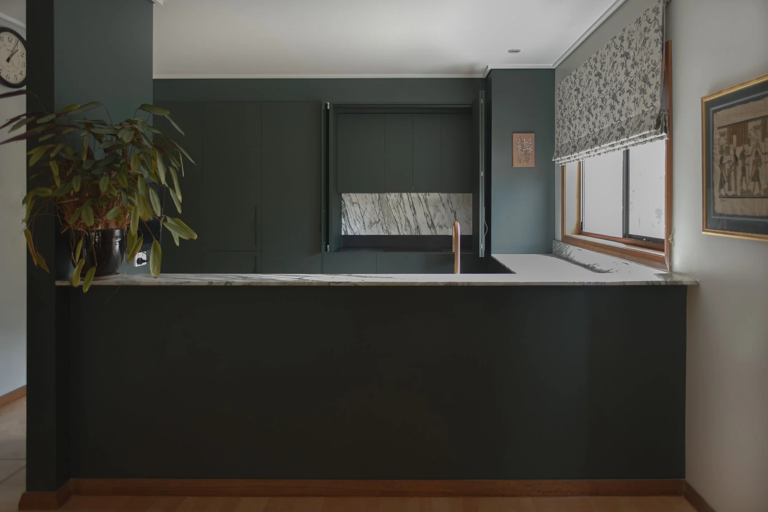 Room with green and white decor, large window with floral Roman shade, wall art, a potted plant on a counter, marble backsplash, and a clock on adjacent wall.