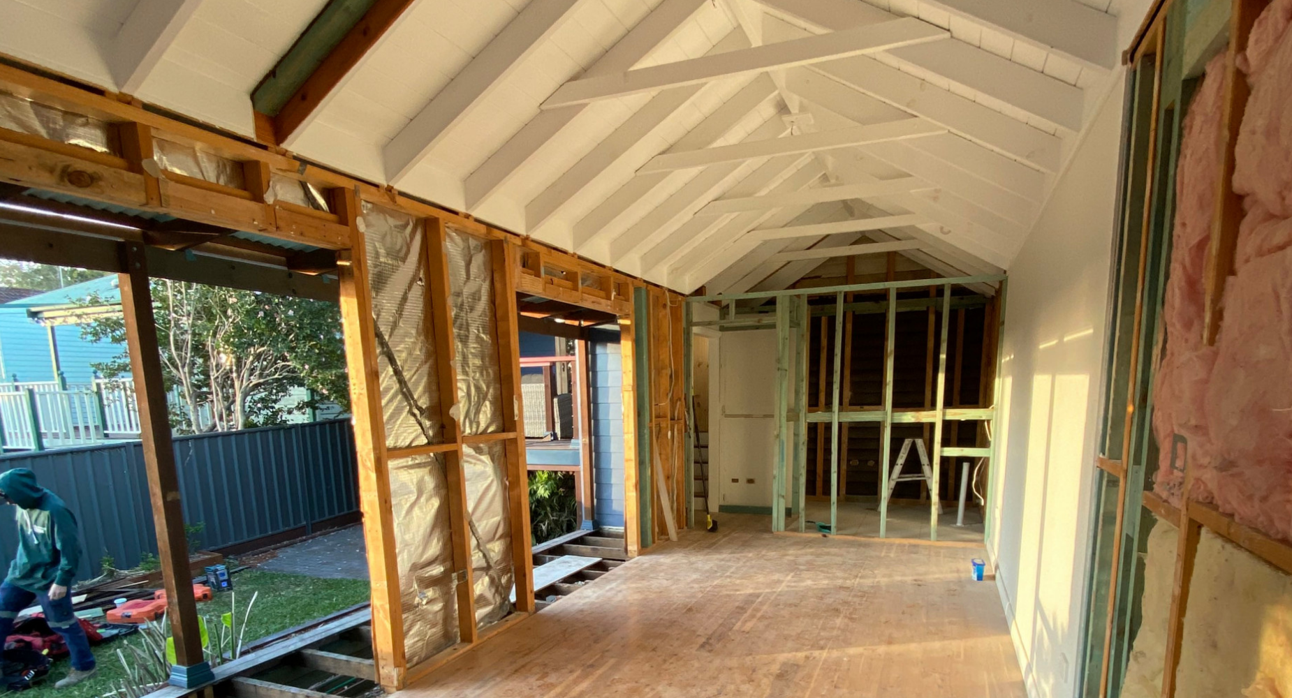 Interior of a house under construction, showing exposed framing and roofing, with a ladder and construction tools visible.