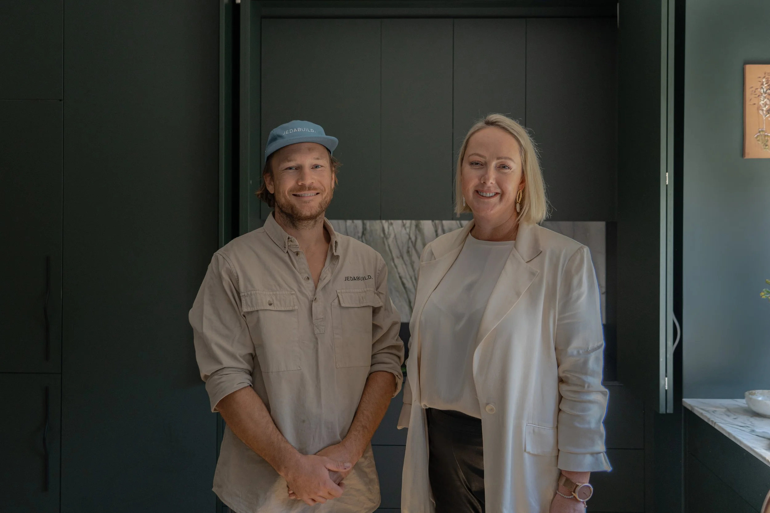 Two people standing side by side indoors, smiling at the camera. The person on the left is a man with a beard, wearing a beige shirt and a blue cap with the text 'JEDABUILD.' The person on the right is a woman with blonde hair, wearing a cream blazer