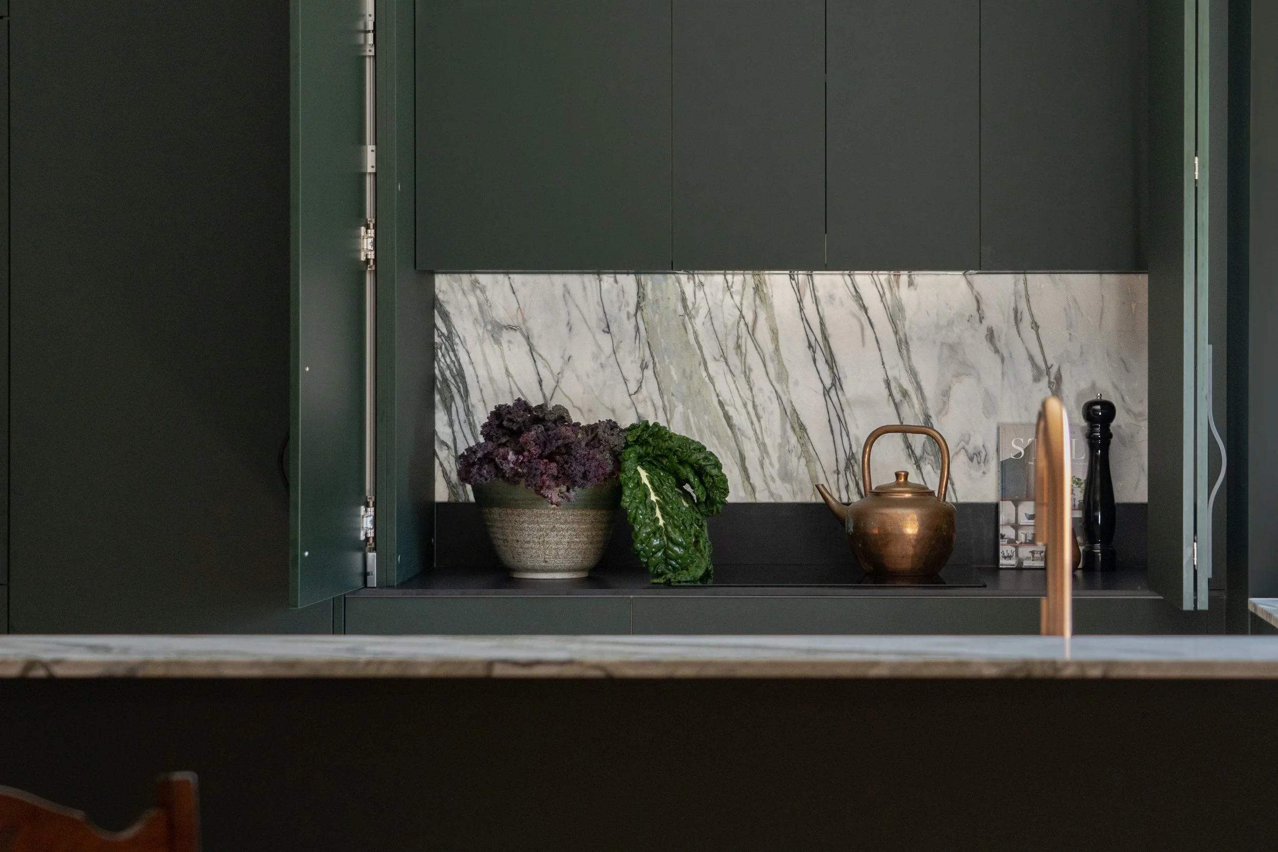 A kitchen cabinet opening to reveal a marble backsplash with purple kale and green leafy vegetables in a bowl, a copper pitcher, a book, and various kitchen utensils.