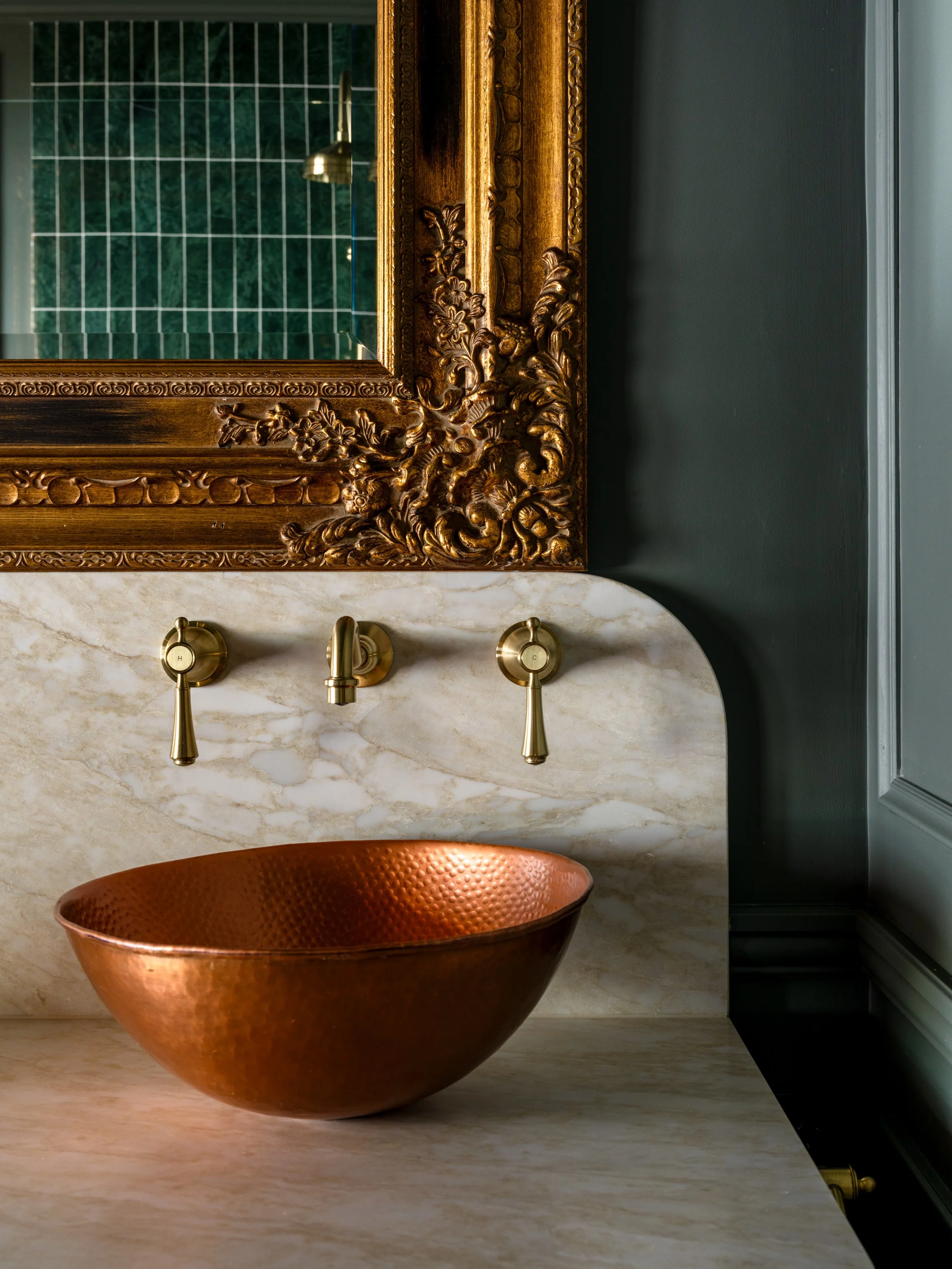 A bathroom sink area with a copper basin, a white marble countertop, and a gold-framed mirror reflecting green tiled walls and a gold lamp.