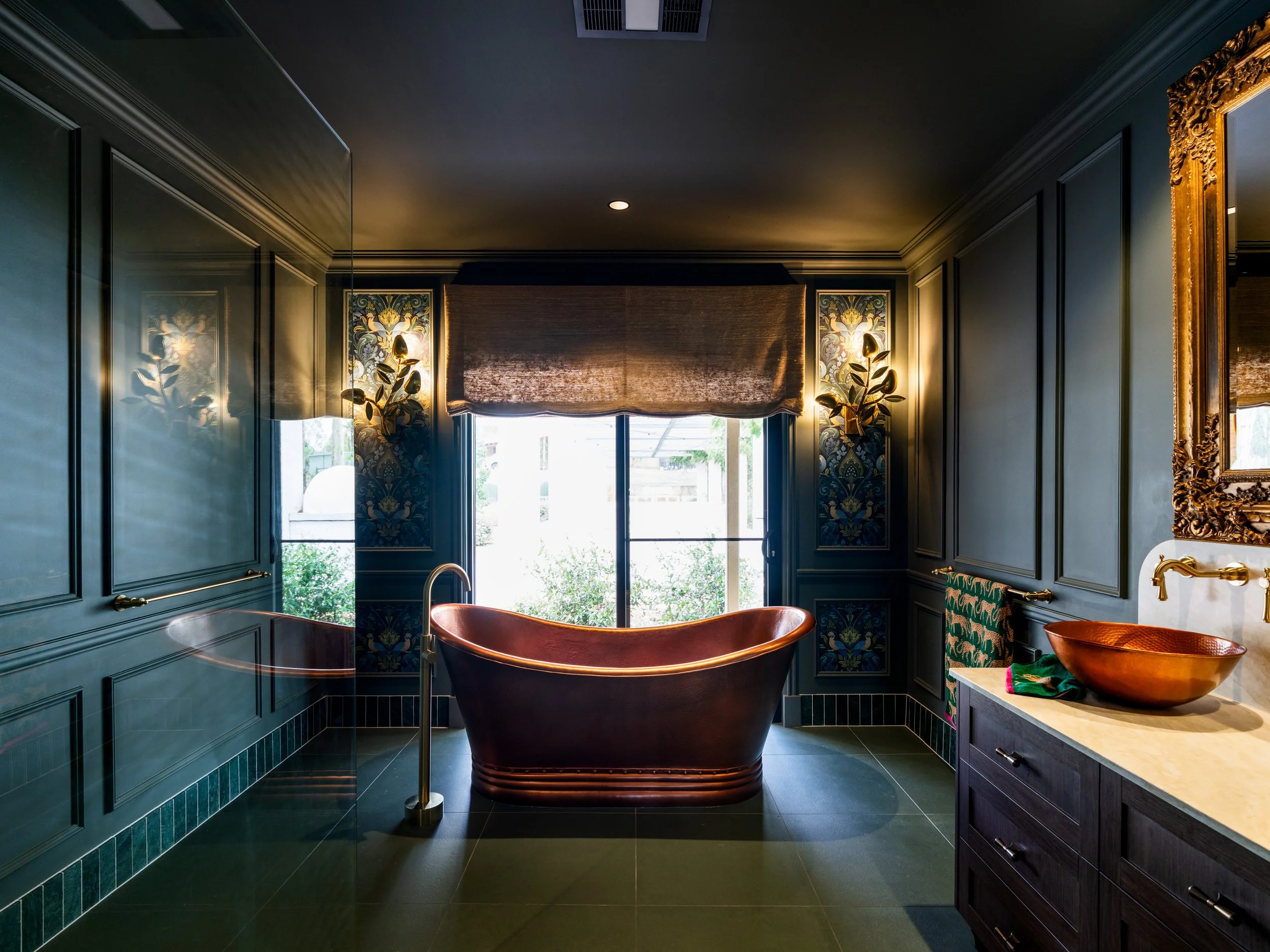A luxurious bathroom with a freestanding bathtub, dark green paneled walls, decorative wallpaper behind the window, and elegant lighting fixtures flanking the window.