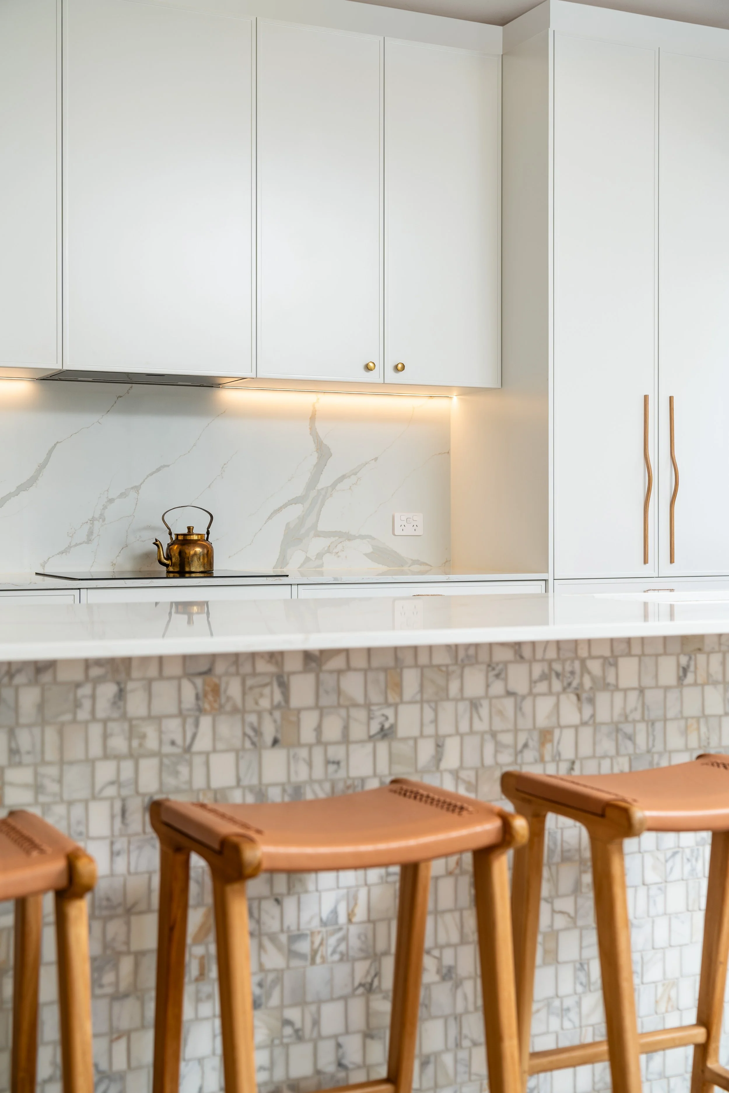 A modern kitchen with white cabinetry, marble backsplash, and a marble countertop with wooden stools in the foreground.