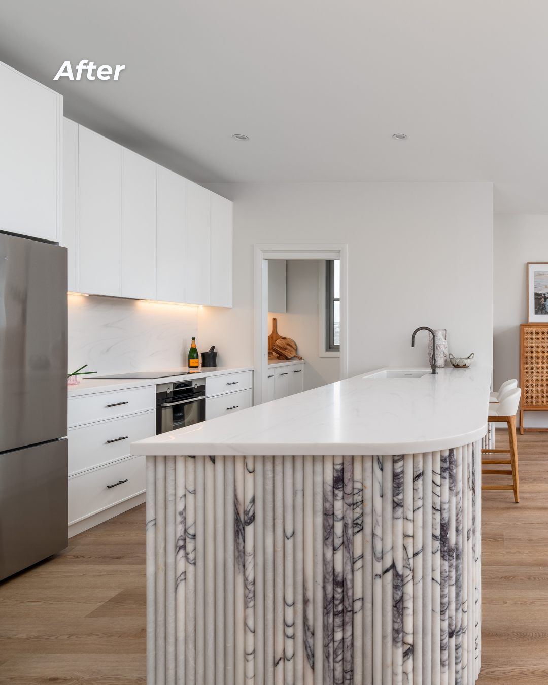 Modern kitchen with white cabinets, marble island, and stainless steel appliances. The kitchen has light wood flooring and minimal decor, with a small doorway leading to another room.