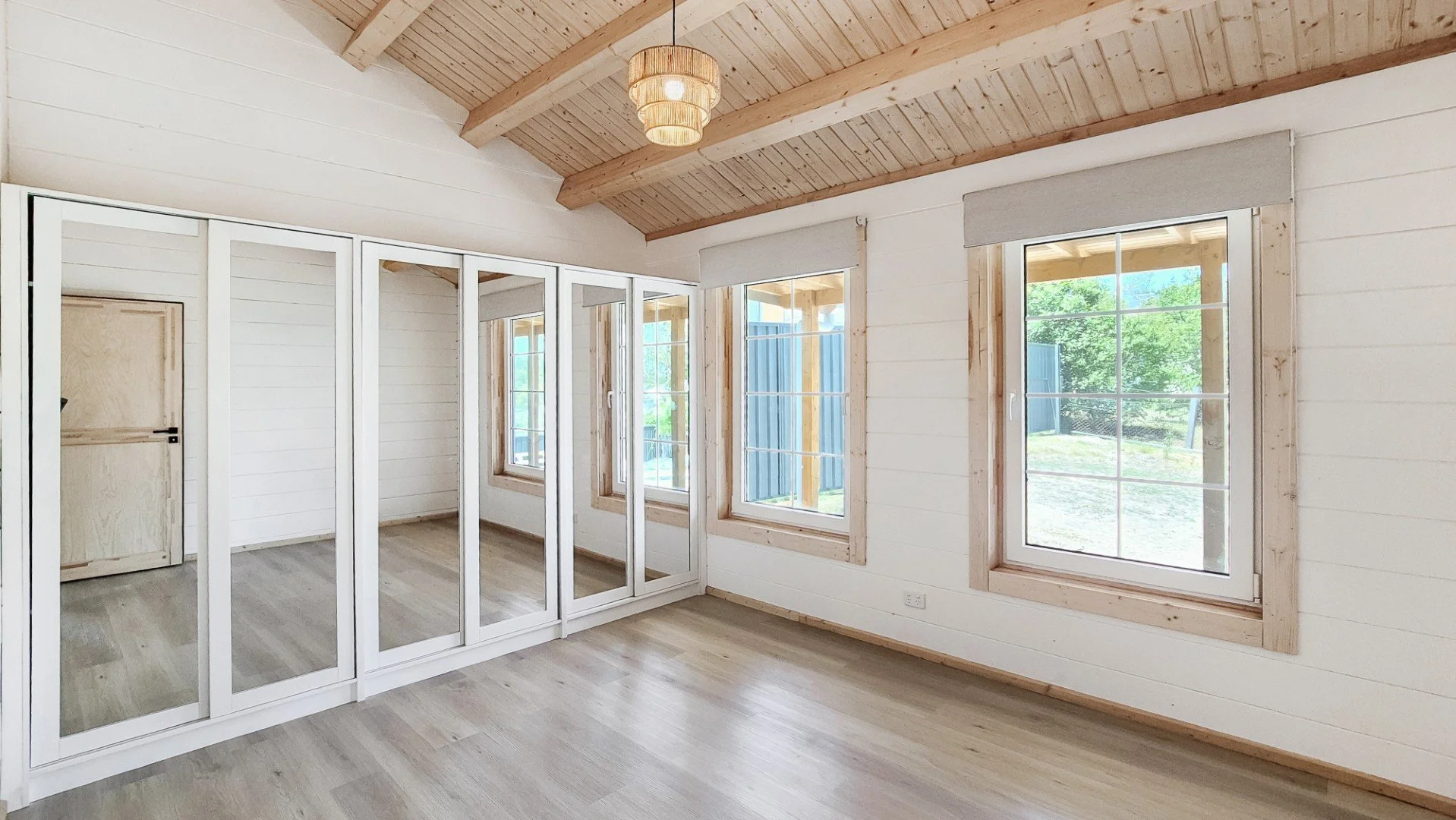 Empty room with light wood floors, white walls, wood-beamed ceiling, large windows with blinds, and a door with a lock.