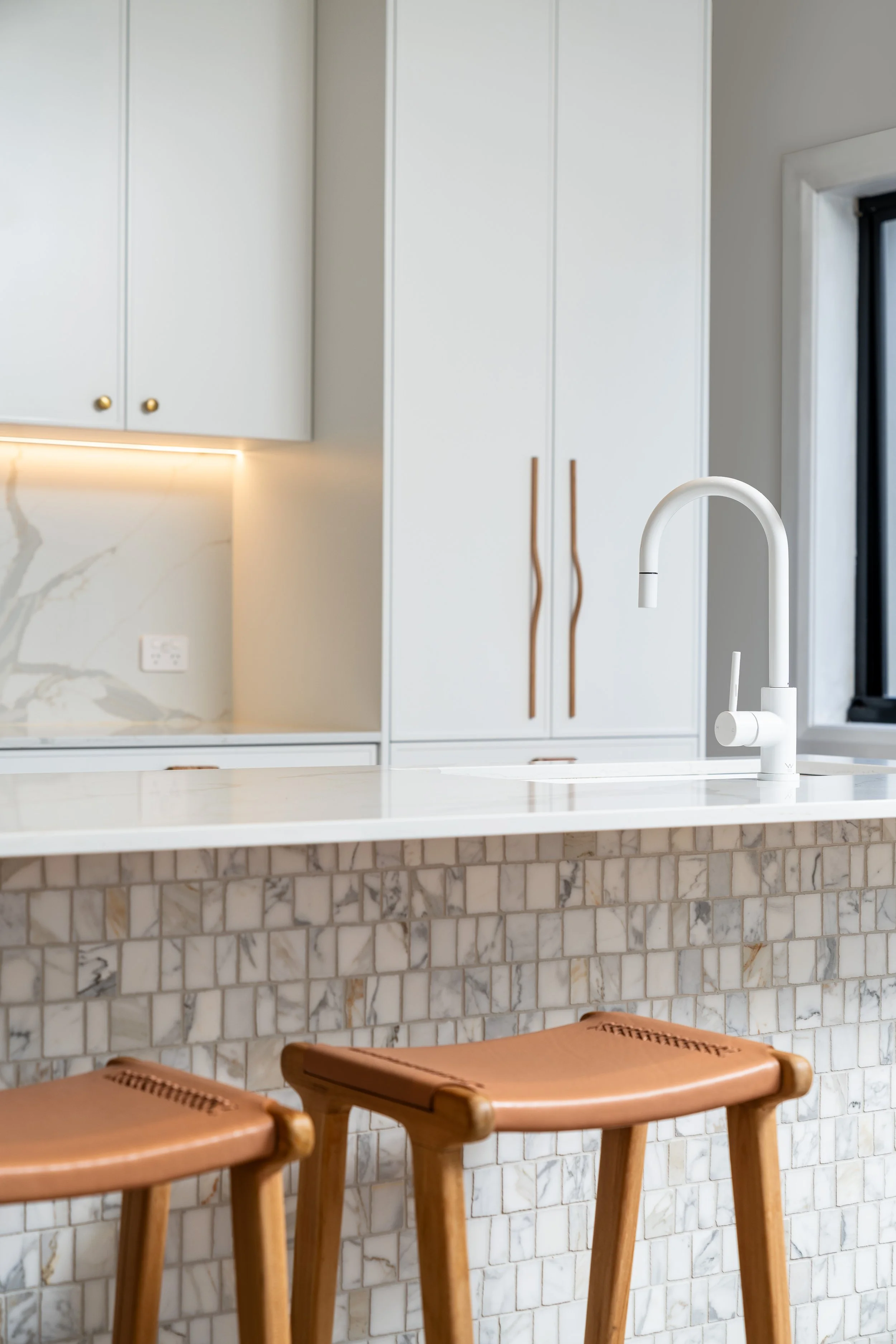 A modern kitchen with white cabinets, a marble backsplash, a white countertop, and wooden stools in the foreground. There is a window on the right side and a faucet over the sink.