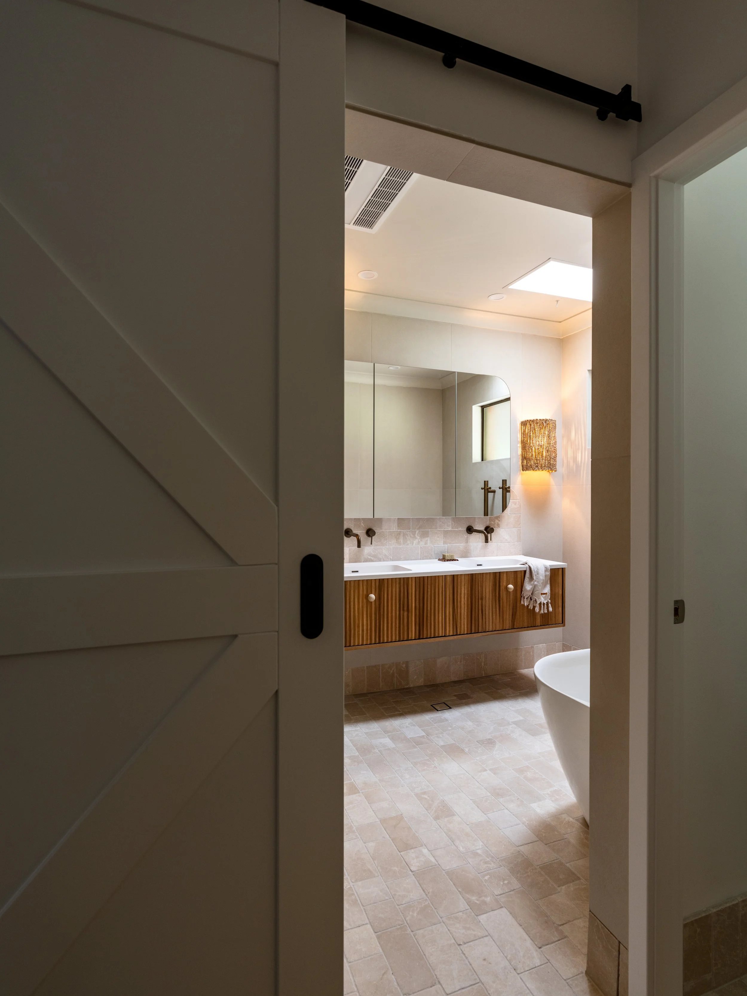 View into a modern bathroom through an open sliding door, featuring a large mirror, a double sink vanity with wooden cabinet, beige tiled walls and floor, and a white toilet.