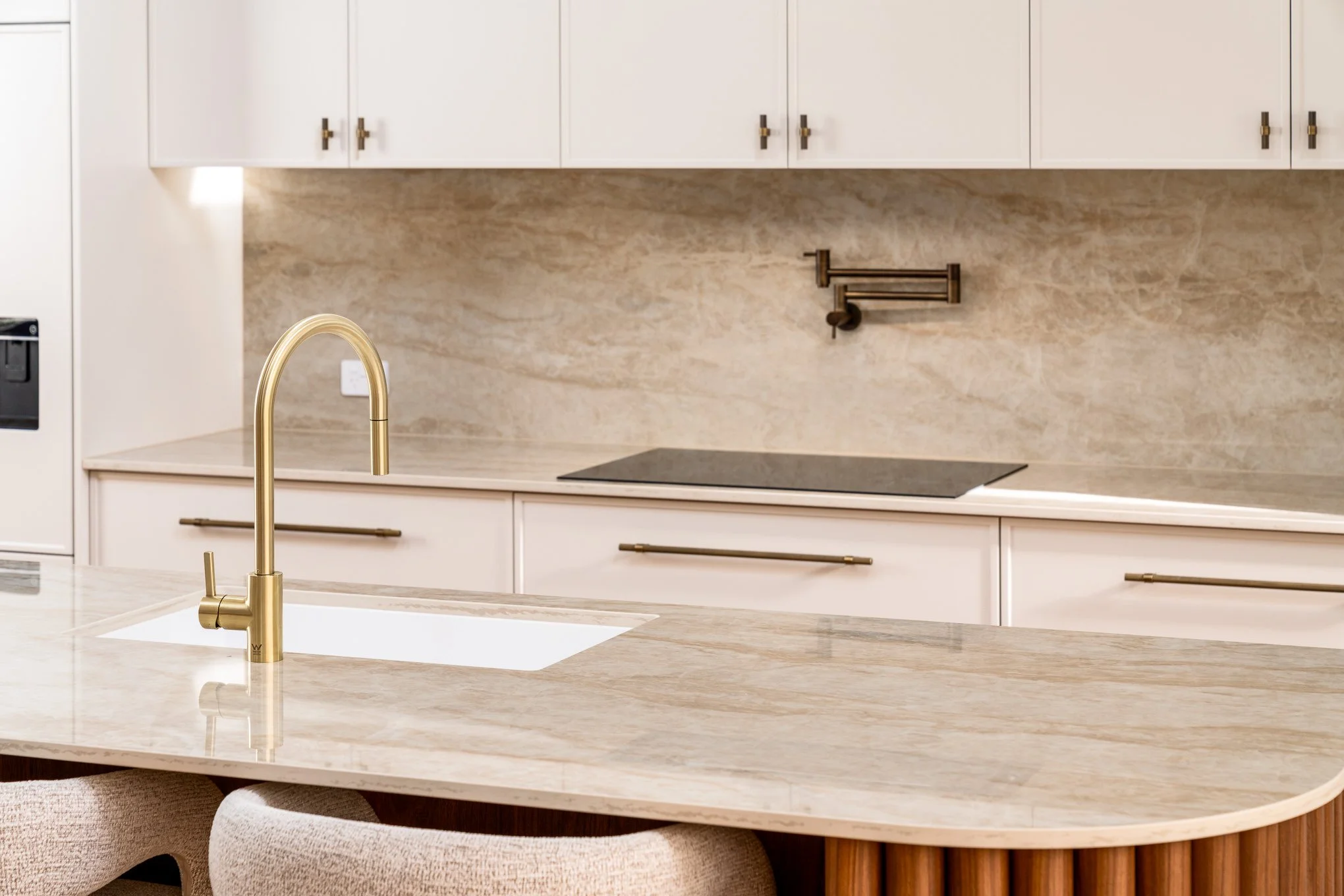 A modern kitchen with a beige marble countertop, a gold faucet, white cabinets with gold hardware, and a beige marble backsplash. A black cooktop is integrated into the countertop, and hanging shelves are mounted on the wall.