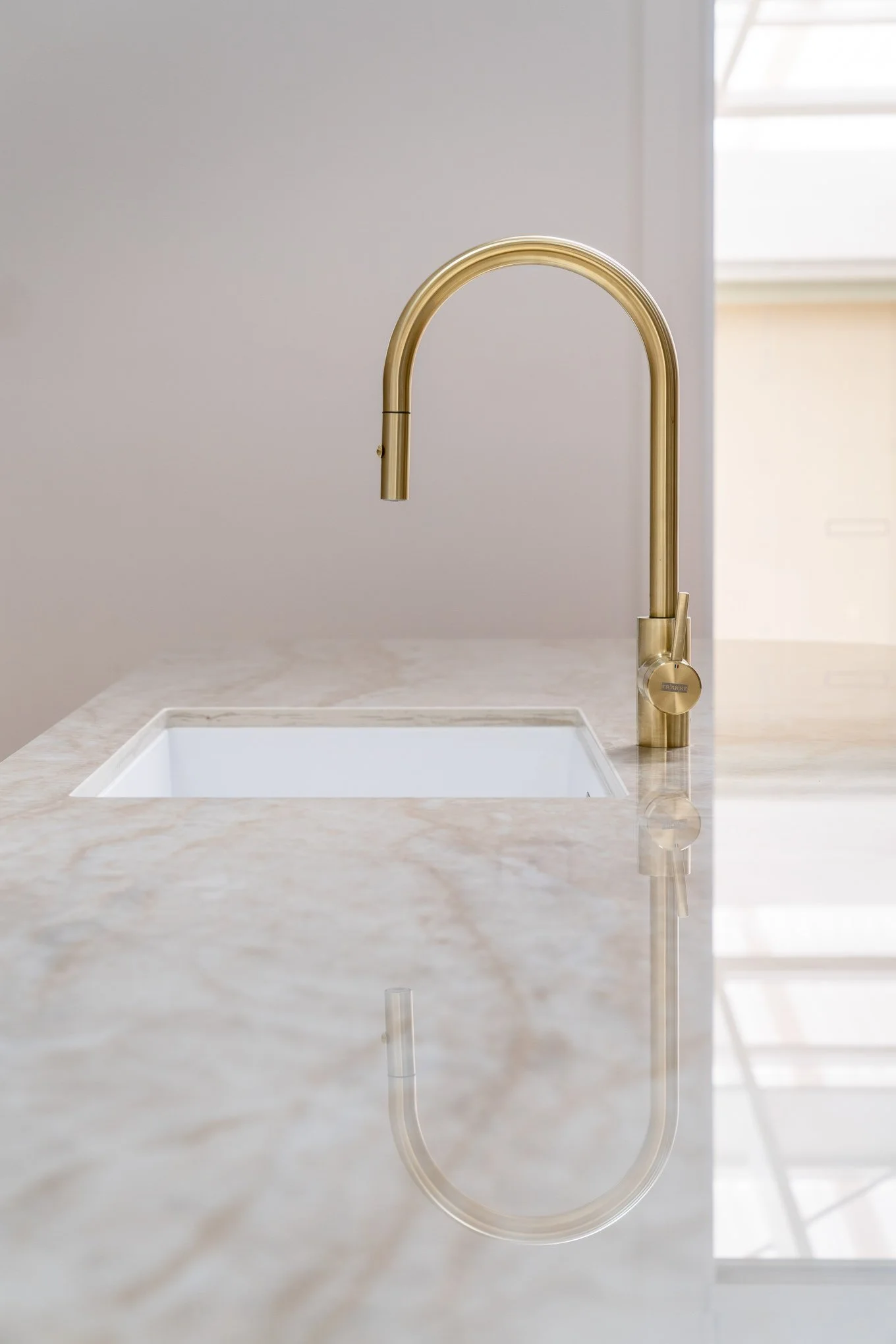 Close-up of a modern gold kitchen faucet mounted on a white marble countertop with a built-in sink, in a bright kitchen setting.
