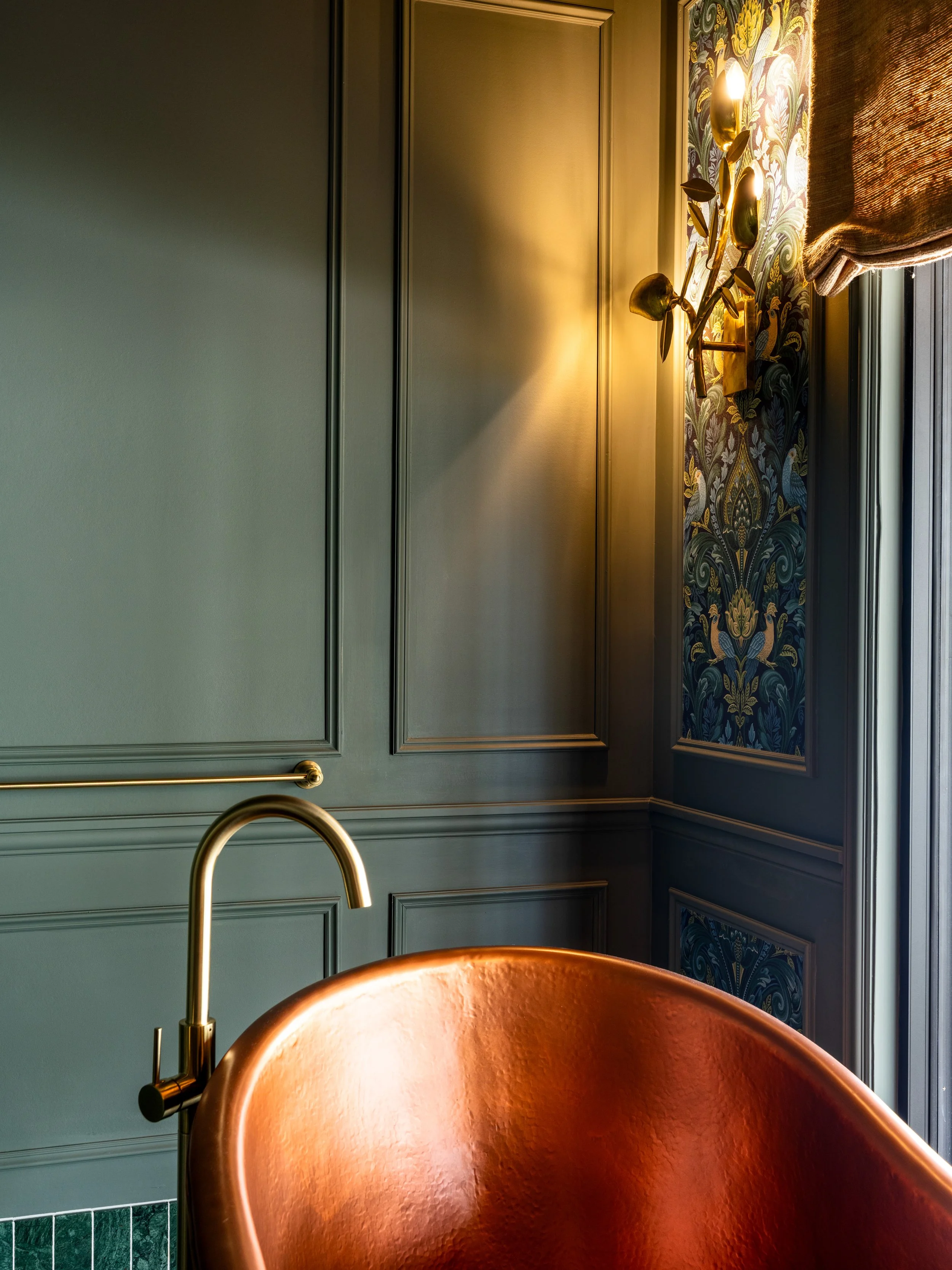 Close-up of a copper bathtub in a bathroom with dark green paneling and patterned wallpaper, featuring a gold-colored faucet and a wall sconce with floral design.