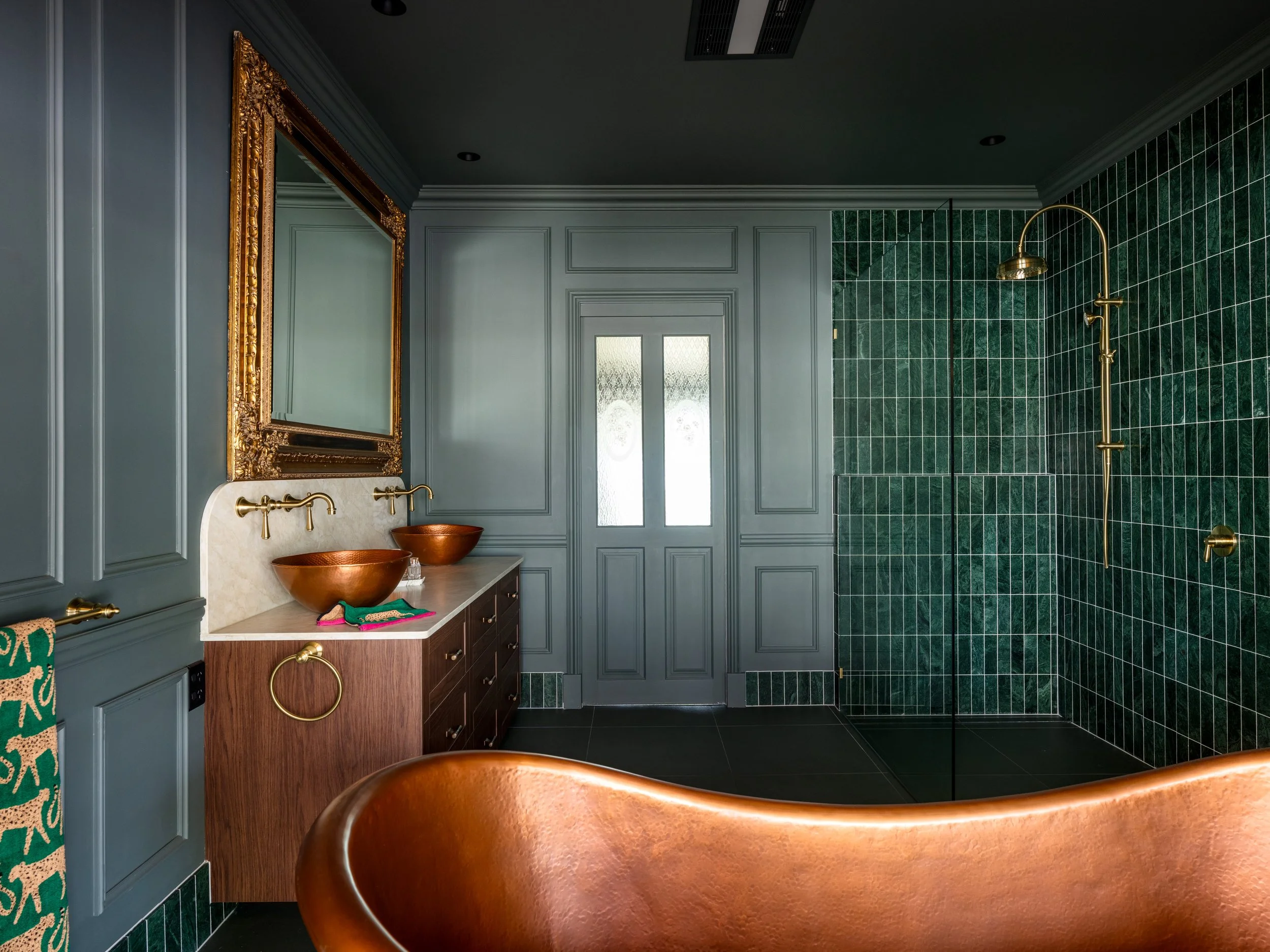 A bathroom featuring charcoal gray paneled walls, green tiled shower walls, vintage brass fixtures, a large ornate gold mirror, and a double copper vessel sink on a wooden vanity.