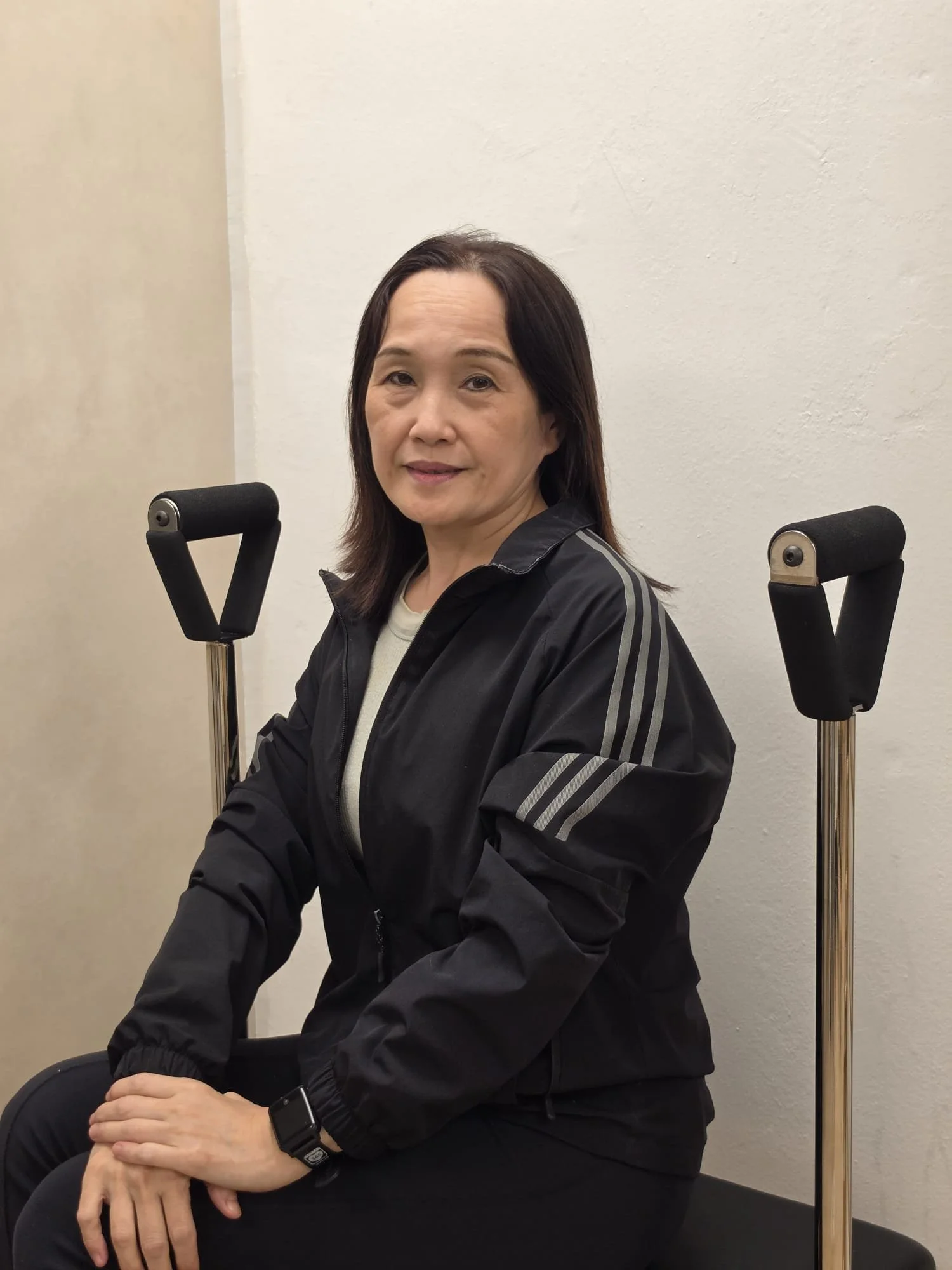 A woman with dark hair wearing a black jacket, sitting on a chair with crutches behind her, against a plain wall.