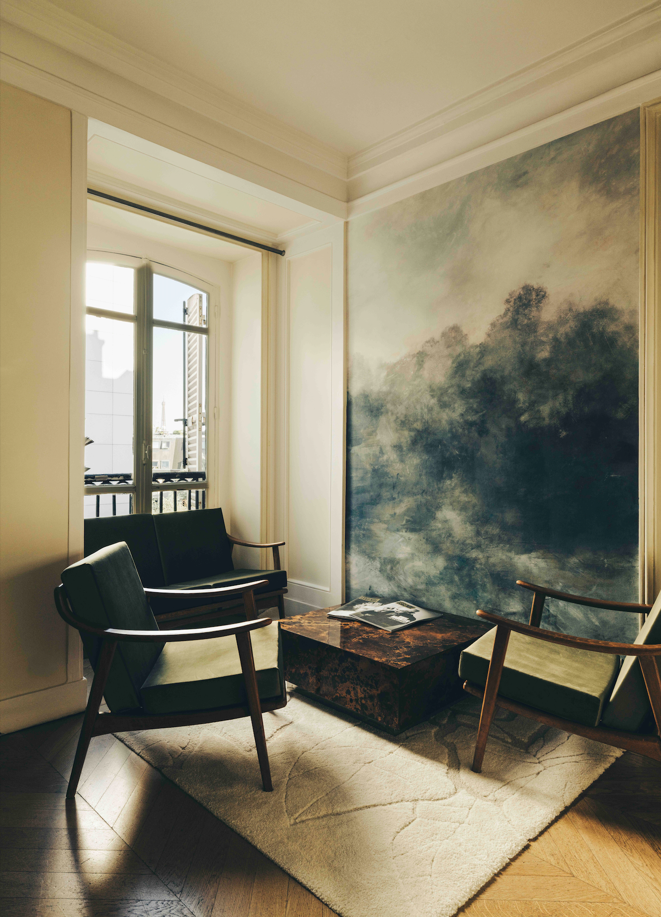 Salon avec deux fauteuils en bois et tissu vert autour d'une table en pierre, mur avec peinture murale évoquant un paysage, fenêtre ouverte laissant entrer la lumière