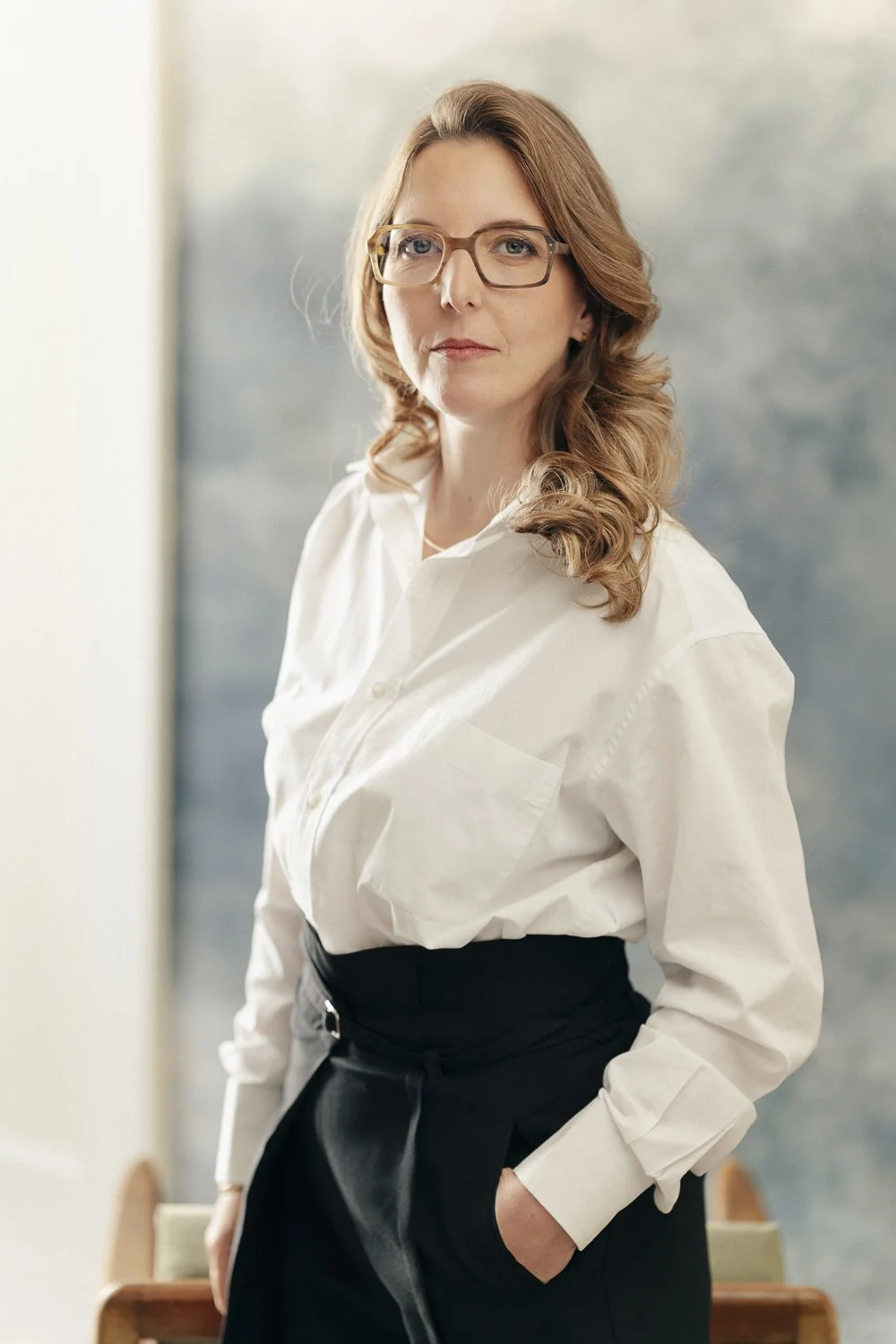 Femme aux cheveux bruns, portant des lunettes, en chemise blanche et pantalon noir, posant avec confiance dans un environnement intérieur.