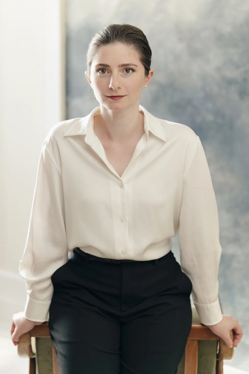 Une femme habillée en chemise blanche et pantalon noir, assise sur une chaise en bois, dans un intérieur lumineux.