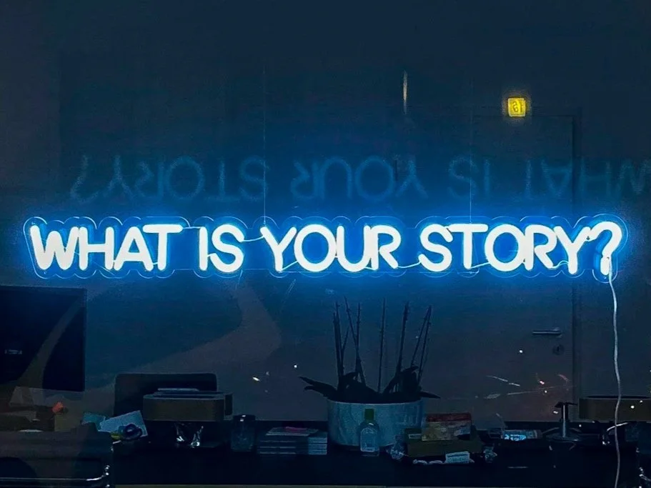 Blue neon sign reading "What is your story?" on a dark wall.