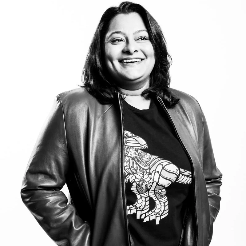 A Black and white photo of a woman smiling, wearing a leather jacket over a graphic t-shirt with a steampunk dinosaur.