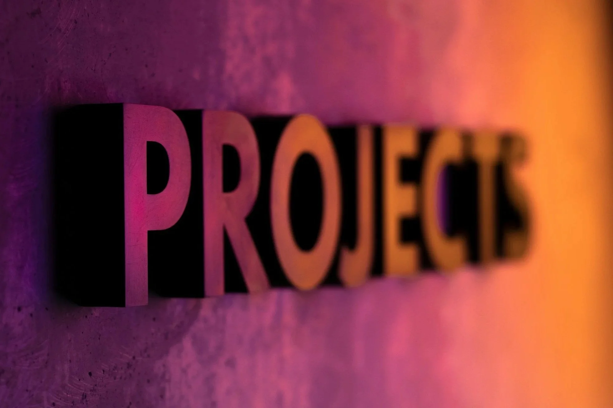 Close-up of black 3D letters spelling 'PROJECTS' on a textured surface, illuminated with pink, purple, and orange lighting.