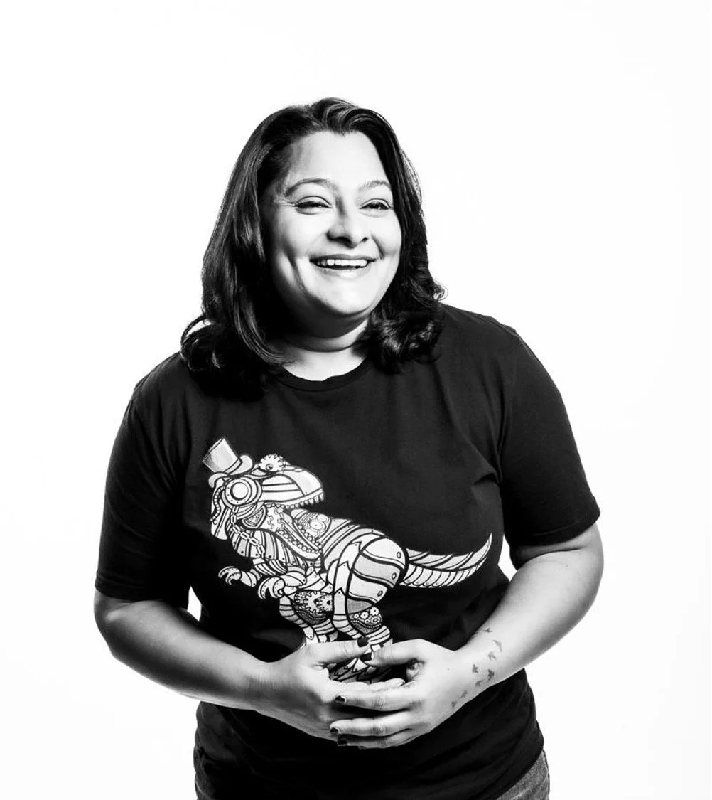 Black and white photo of a woman with shoulder-length hair smiling, wearing a t-shirt with a detailed graphic of a steampunk dinosaur.