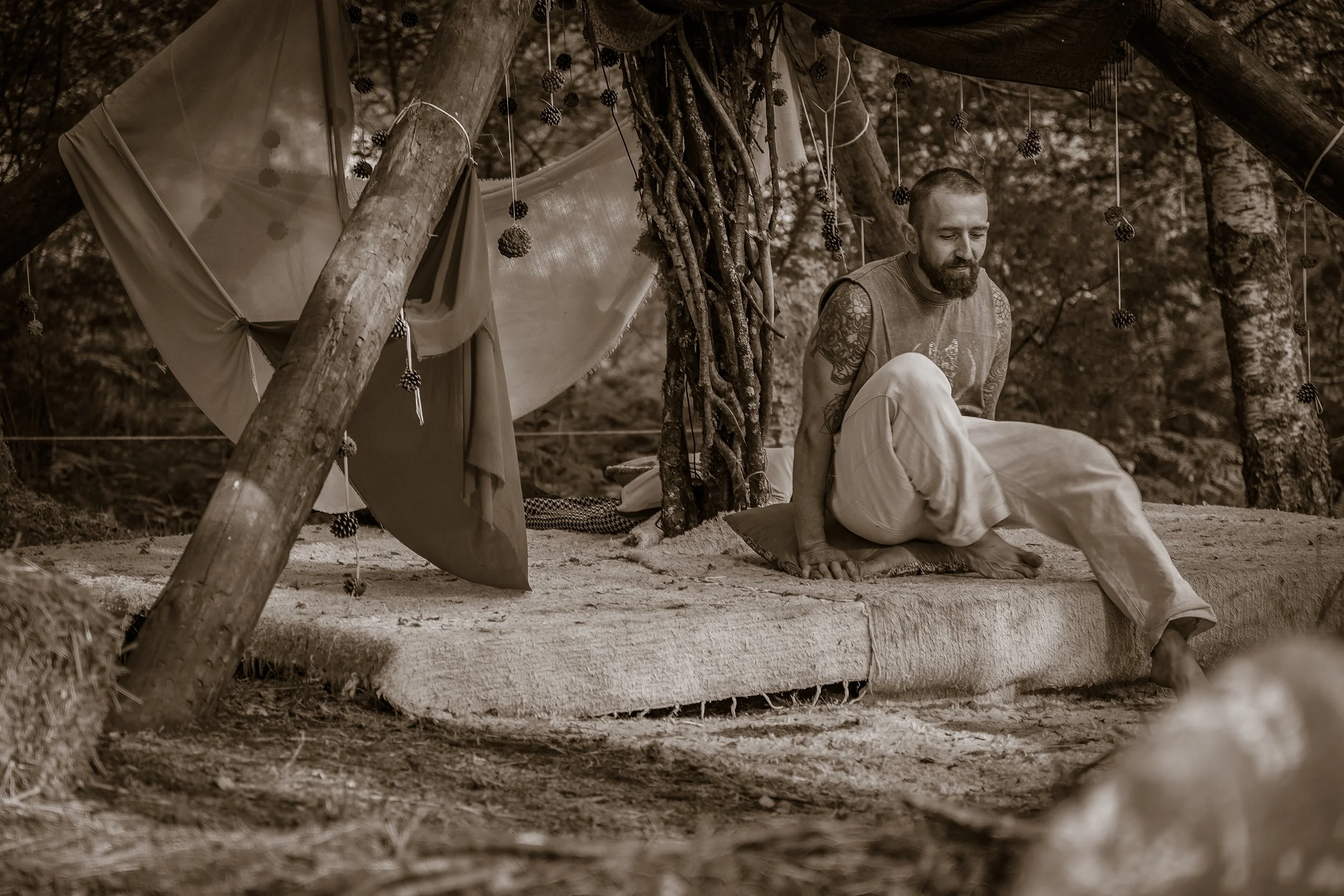 A man with a beard and tattoos sitting on a blanket in a forest, under a makeshift shelter decorated with pinecones and fabric.