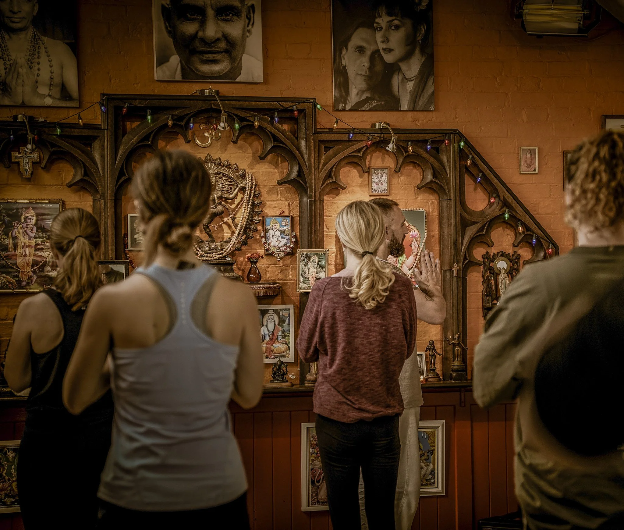People gathered in a room with religious and celebrity portraits on the wall, some with hands in prayer position, engaging in a spiritual or cultural event.