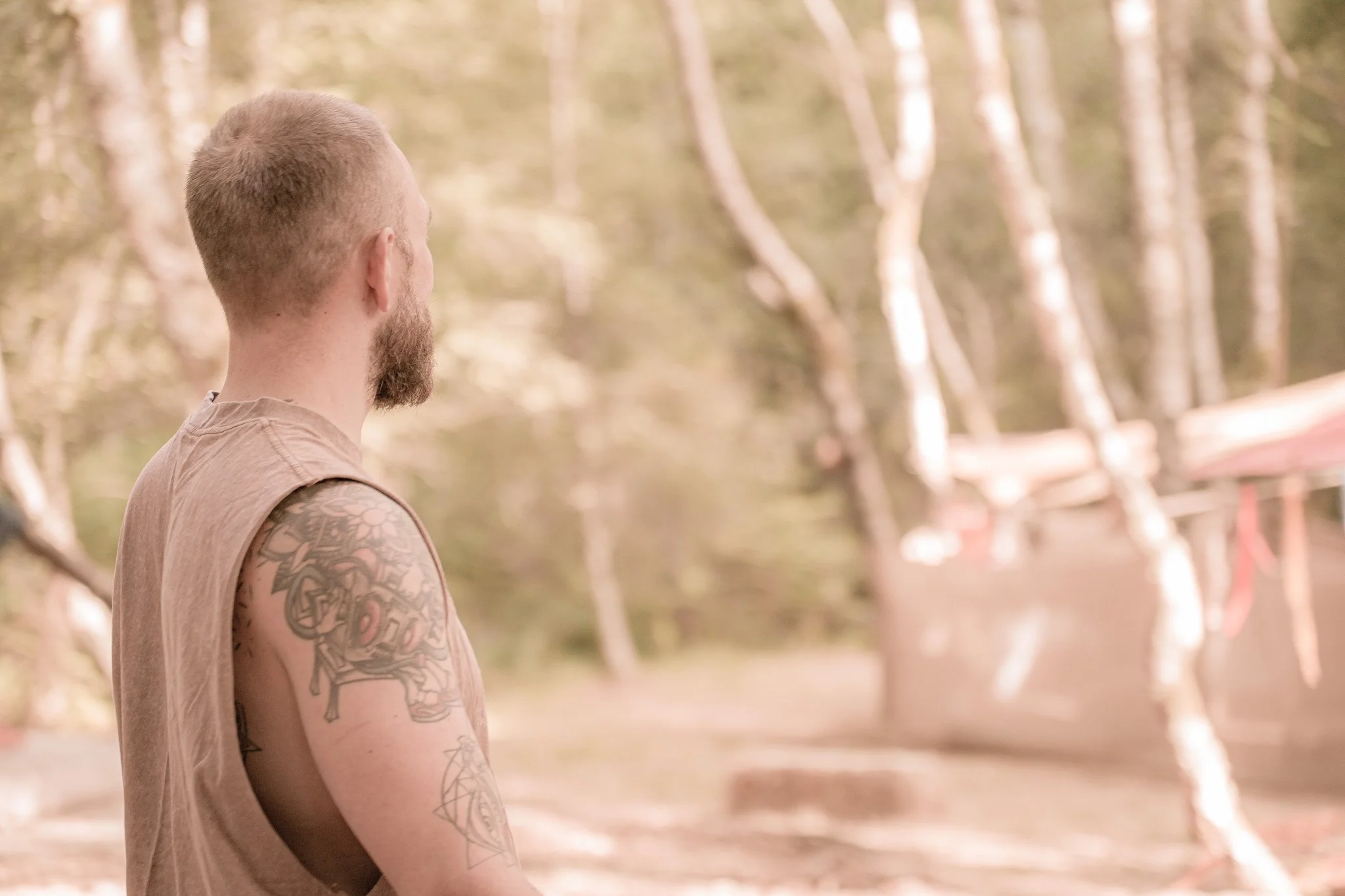 A man with tattoos on his arms and a beard, standing outdoors near a body of water, looking away from the camera.