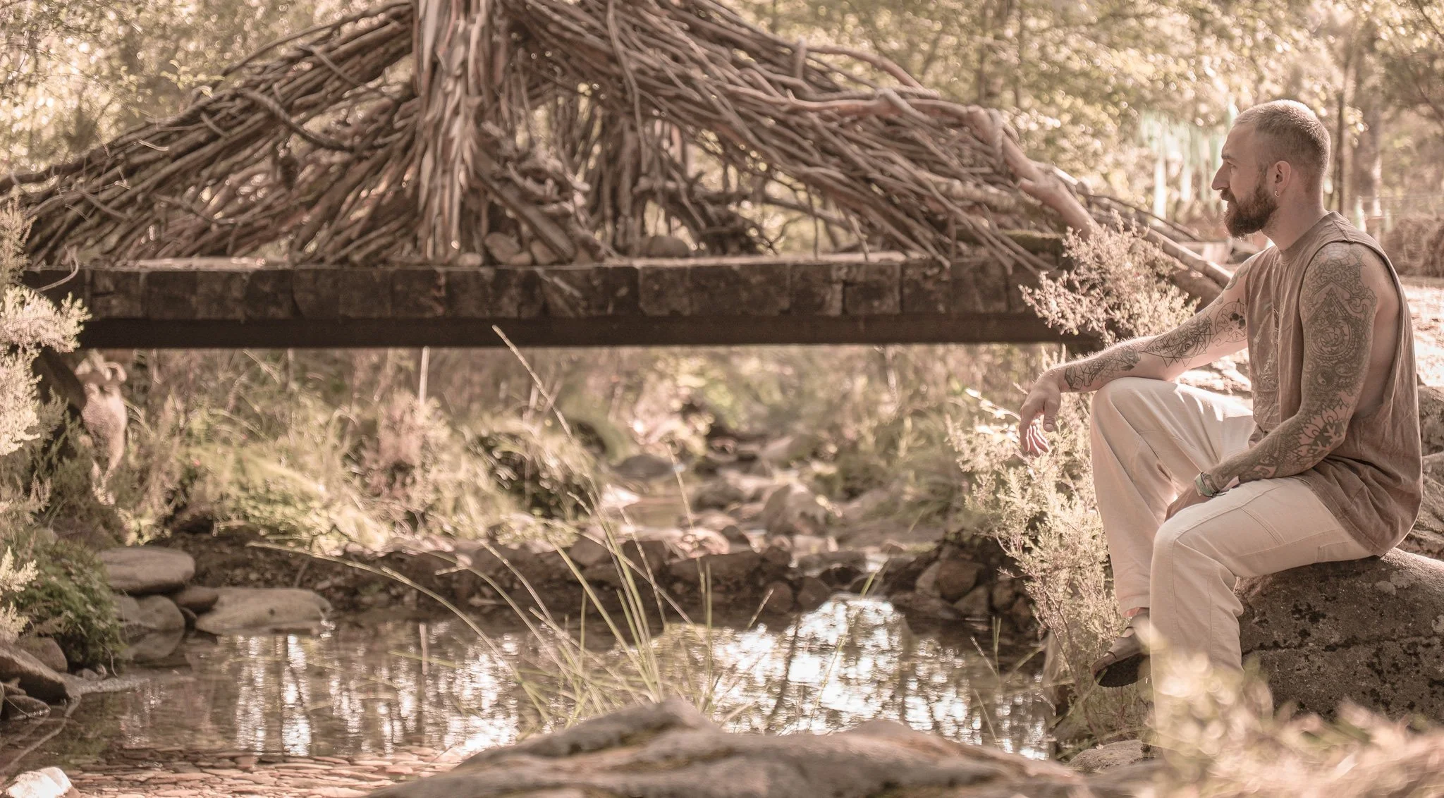 A man with tattoos sitting next to a stream in a wooded area, looking at a makeshift shelter made of branches and logs.