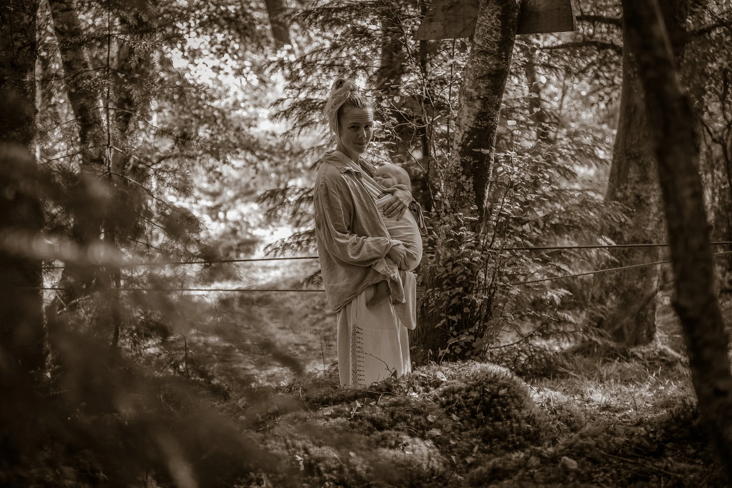 A woman with a child in a forest, the woman holding the child close to her chest and looking at the camera, trees and foliage surround them in a sepia-toned photograph.