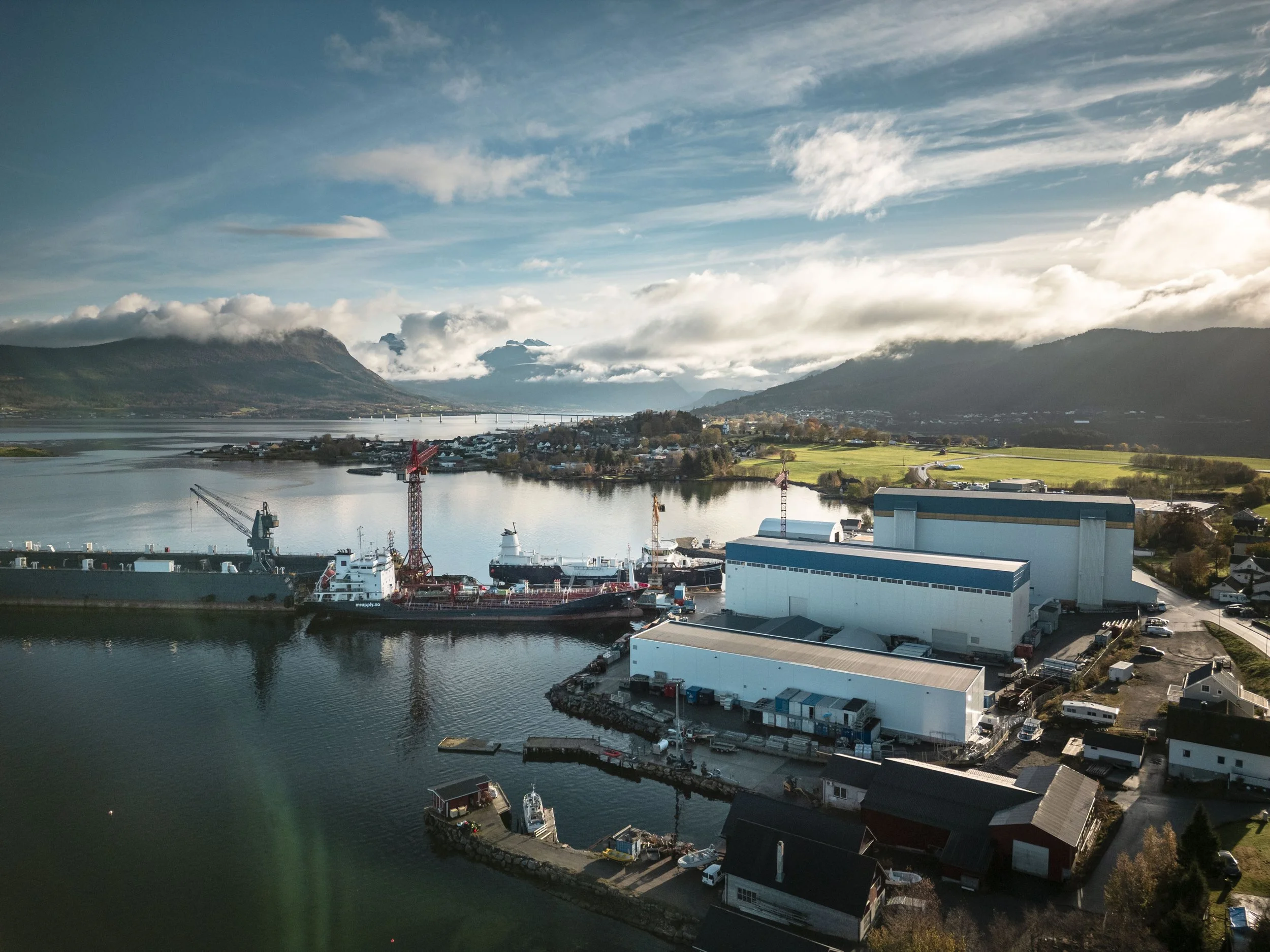 Oversiktsbilde fra Aas Mek sitt verksted på Vestnes. Flere skip ligger inne i havna, og det er utsikt mot Vestnes, Tresfjordbrua og fjordene og fjellene bak. Det er fint vær og lett overskyet