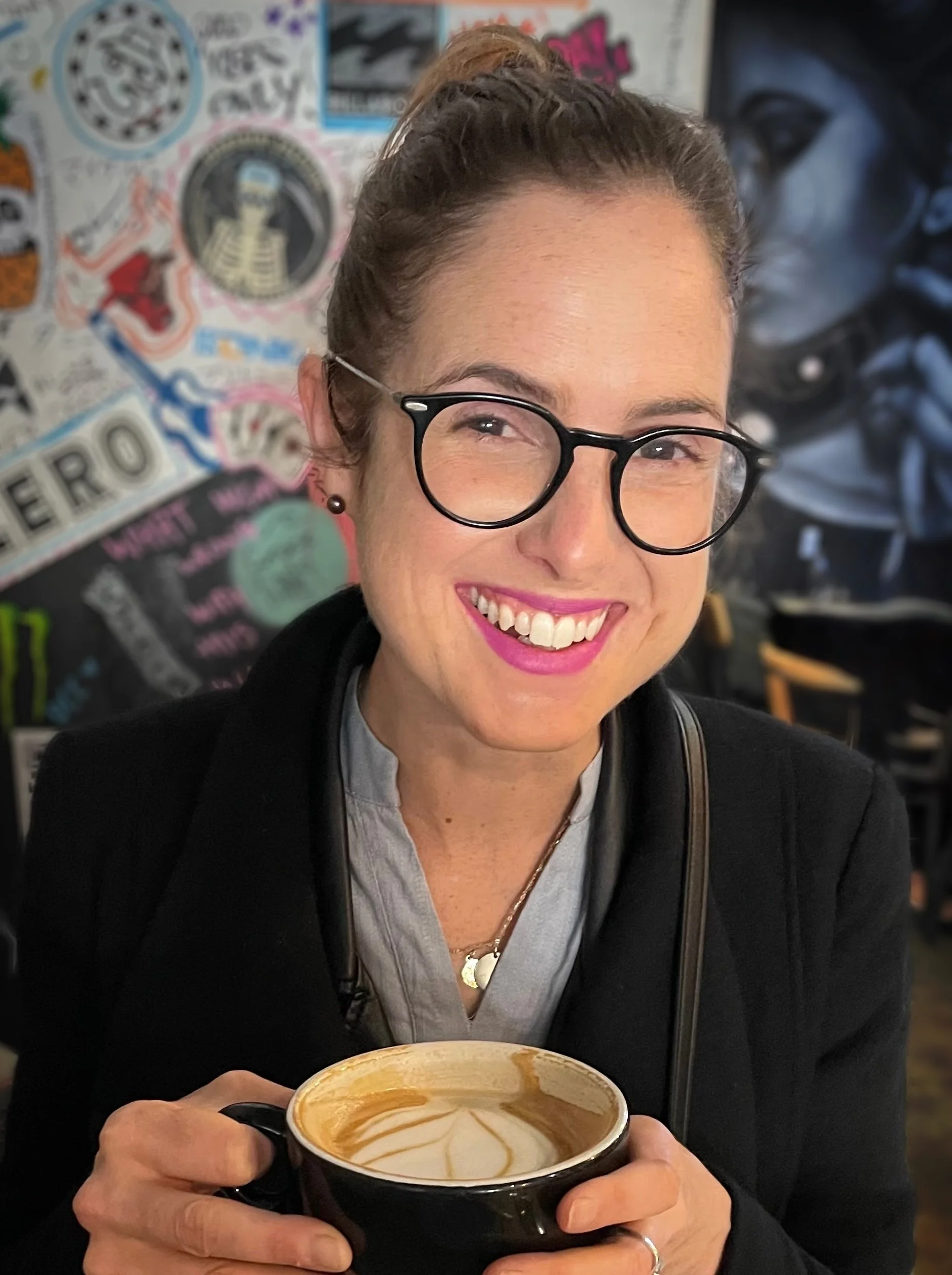 A woman smiling and holding a cup of coffee with latte art in a cafe. She is wearing glasses, earrings, and a dark jacket. There are colorful posters and a painting of a woman in the background.