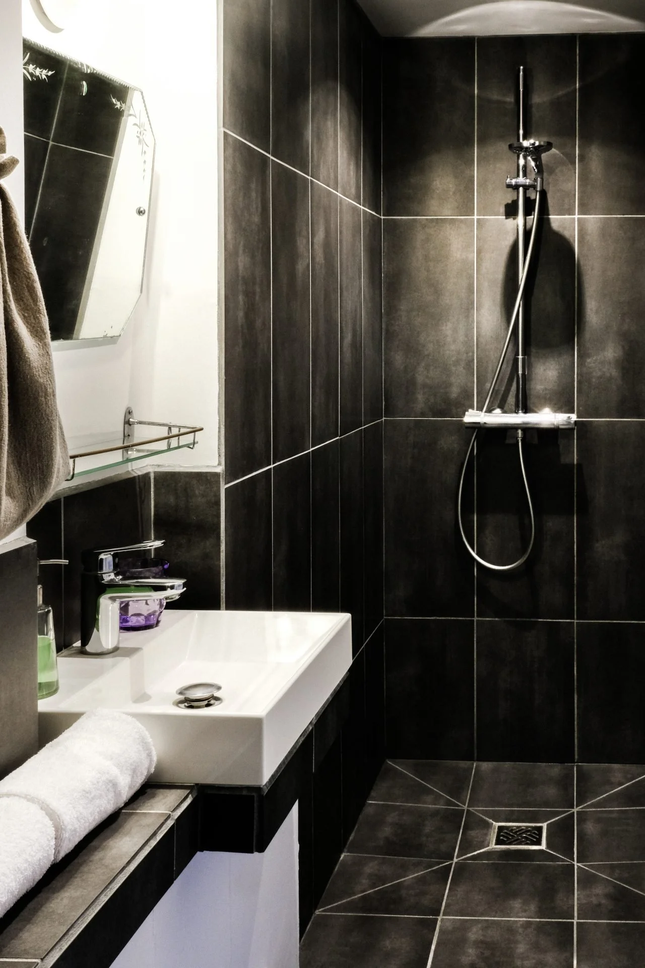 A modern bathroom with dark tiled walls and floor, a white rectangular sink, a mirror, a towel, and a shower head with a hose.