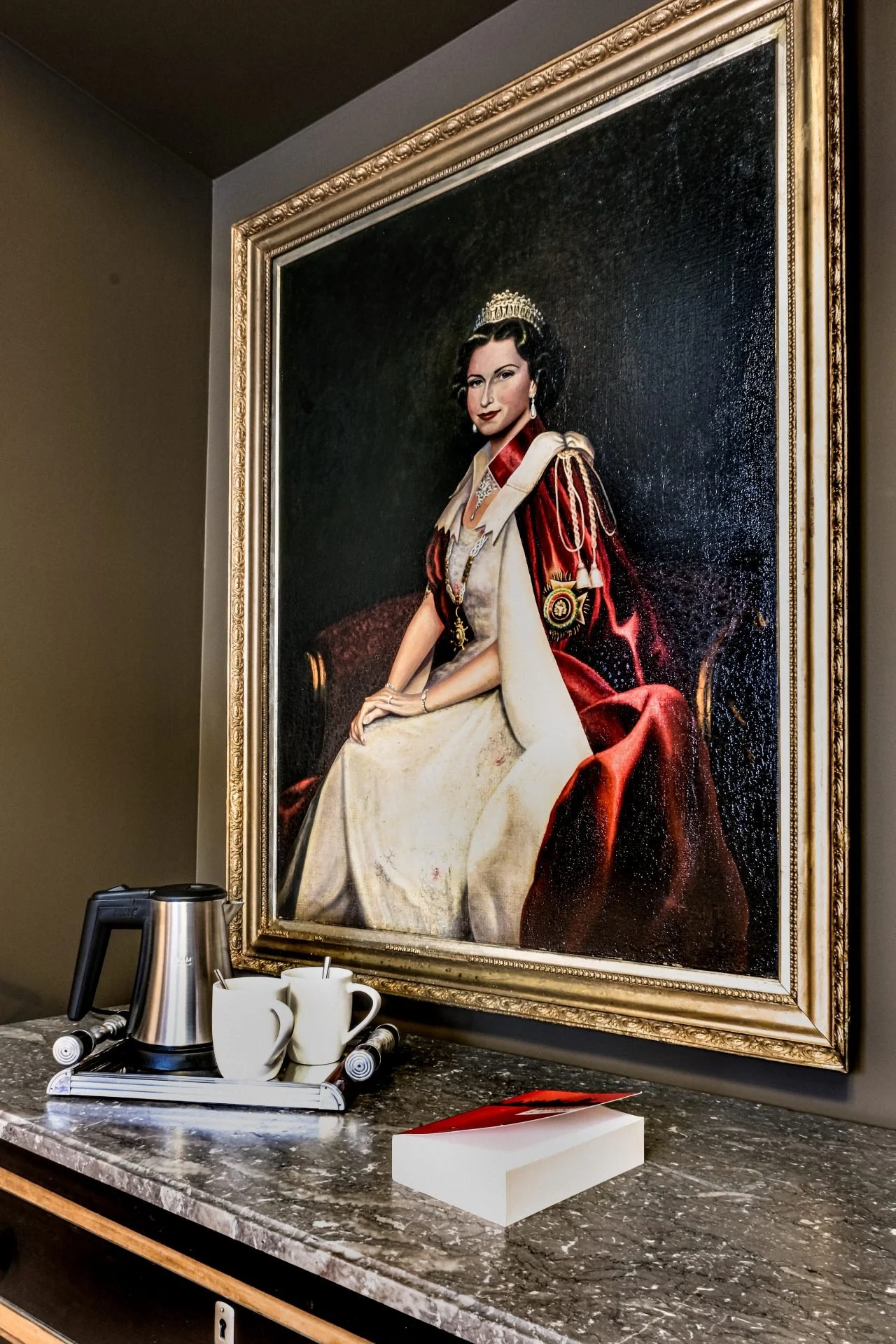 A large framed portrait of a woman dressed in regal attire hangs on a wall. In front of it, on a marble countertop, there is a tray with a coffee pot, cups, and some utensils, along with a red and white book.