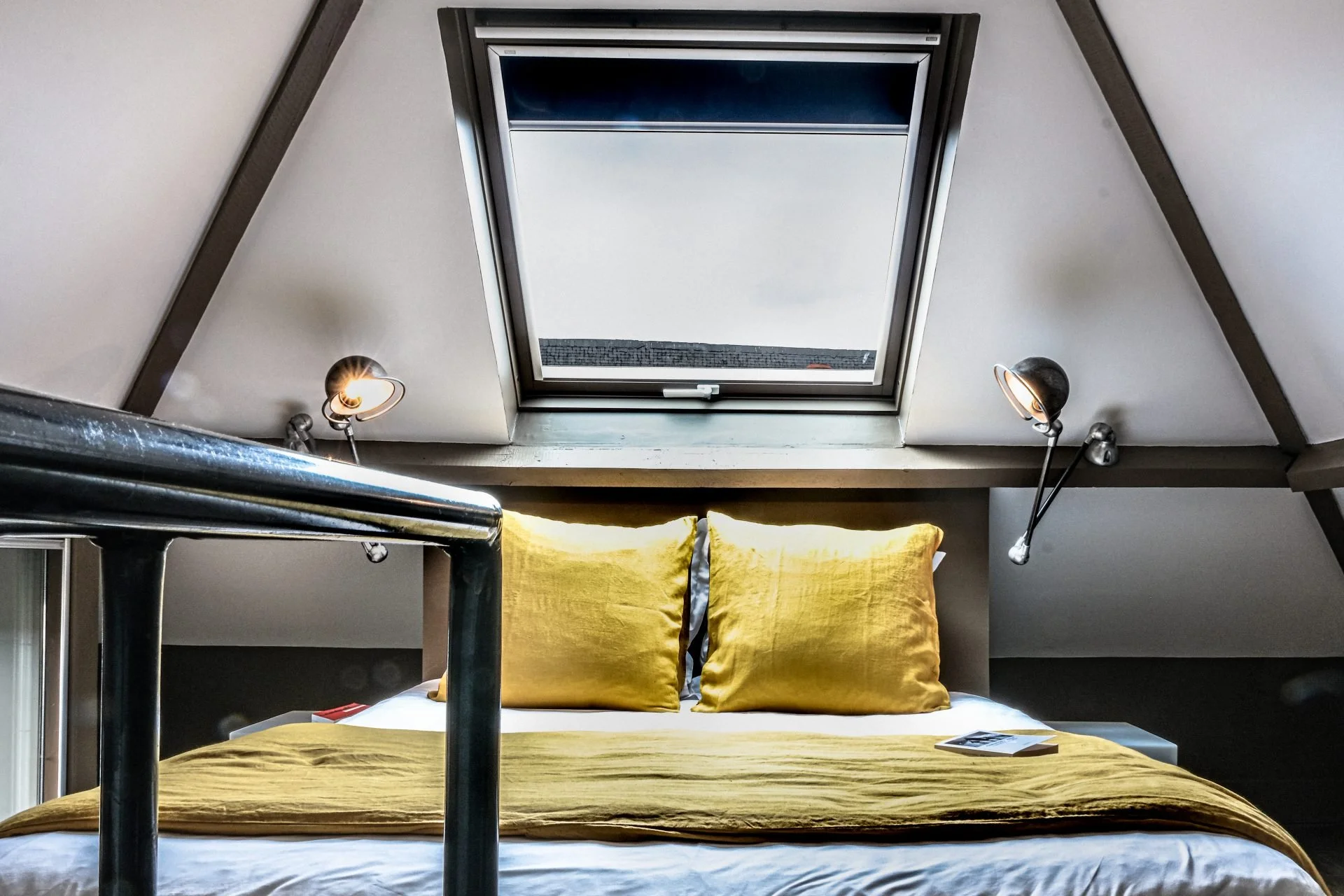 A cozy attic bedroom with a skylight window, two angled reading lamps attached to the wall, and a bed with yellow bedding and pillows. There is a magazine or book on the bed.