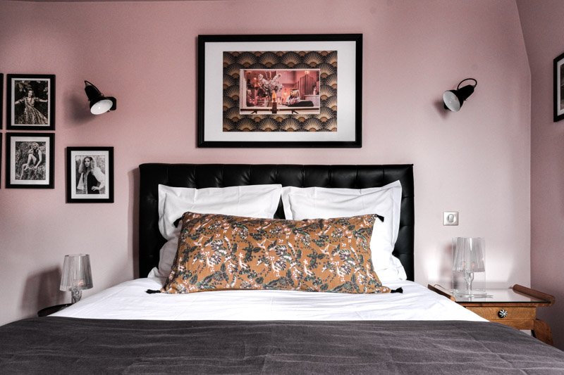 A bedroom with a pink wall, a black leather headboard, white bedding, and a decorative pillow with floral pattern. There are two glass lamps on wooden nightstands, black and white framed photos, and a large framed picture hanging above the bed.