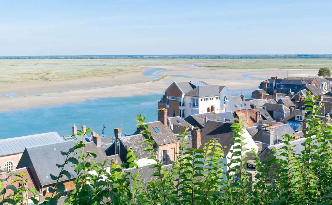 A view of a riverside town with houses in the foreground, overlooking a river and open landscape under a clear sky.