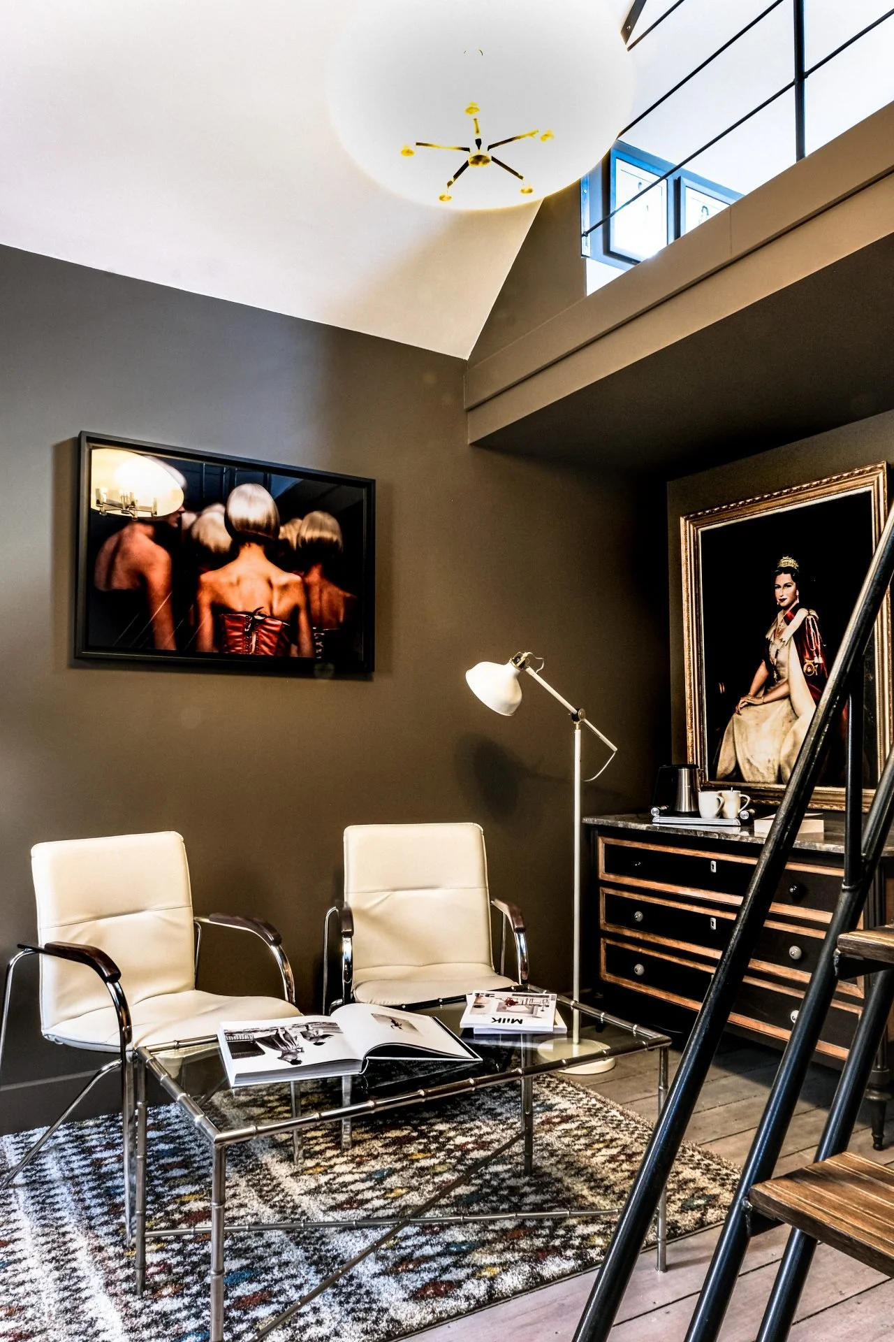 Interior of a modern living space with two white chairs, a glass coffee table with magazines, framed art on the wall, and a patterned rug on wooden flooring.