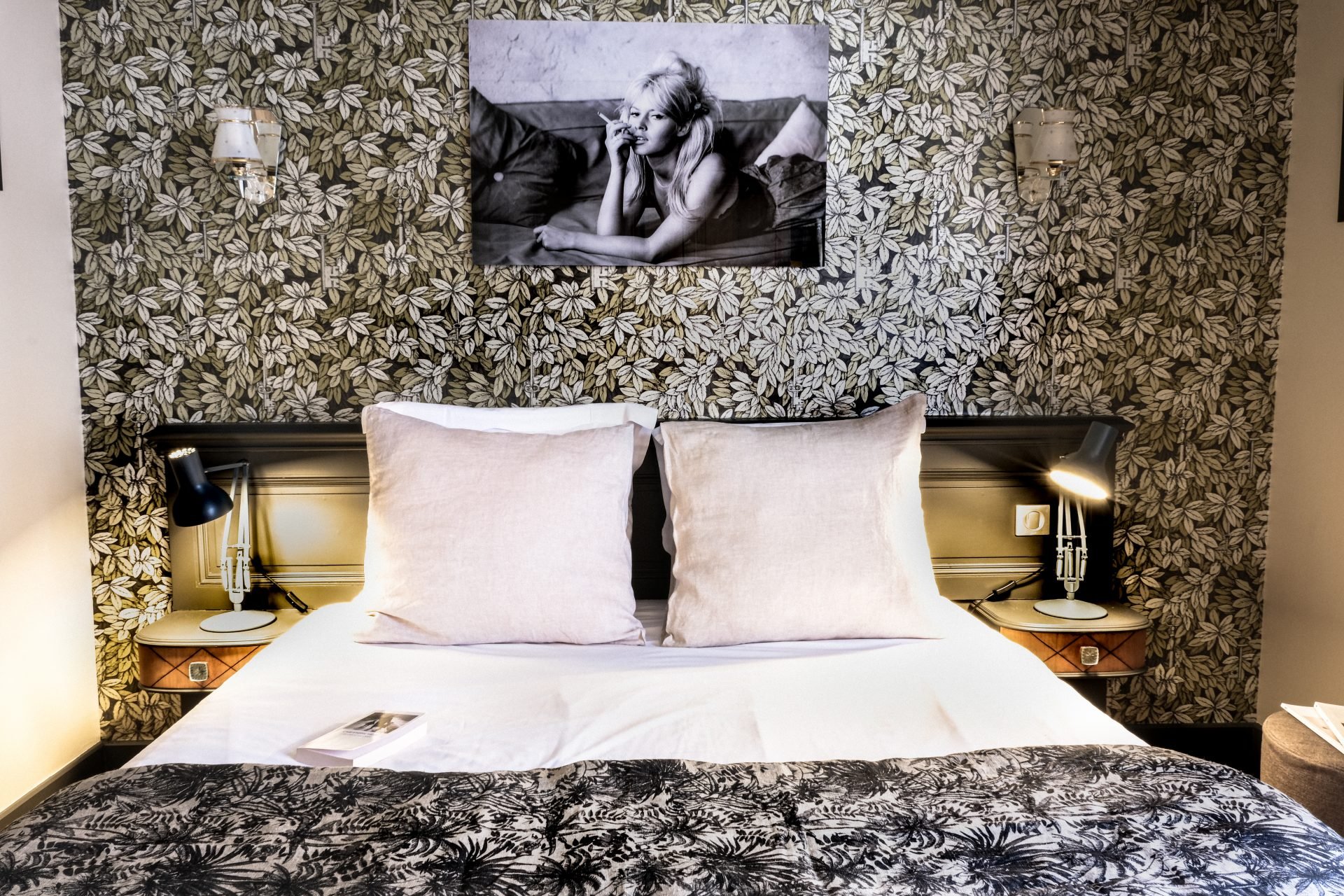 Une chambre à coucher avec du papier peint à motif floral, une photo en noir et blanc d'une femme suspendue au-dessus du lit, deux oreillers blancs, des tables de chevet avec des lampes réglables noires et un livre sur le lit.