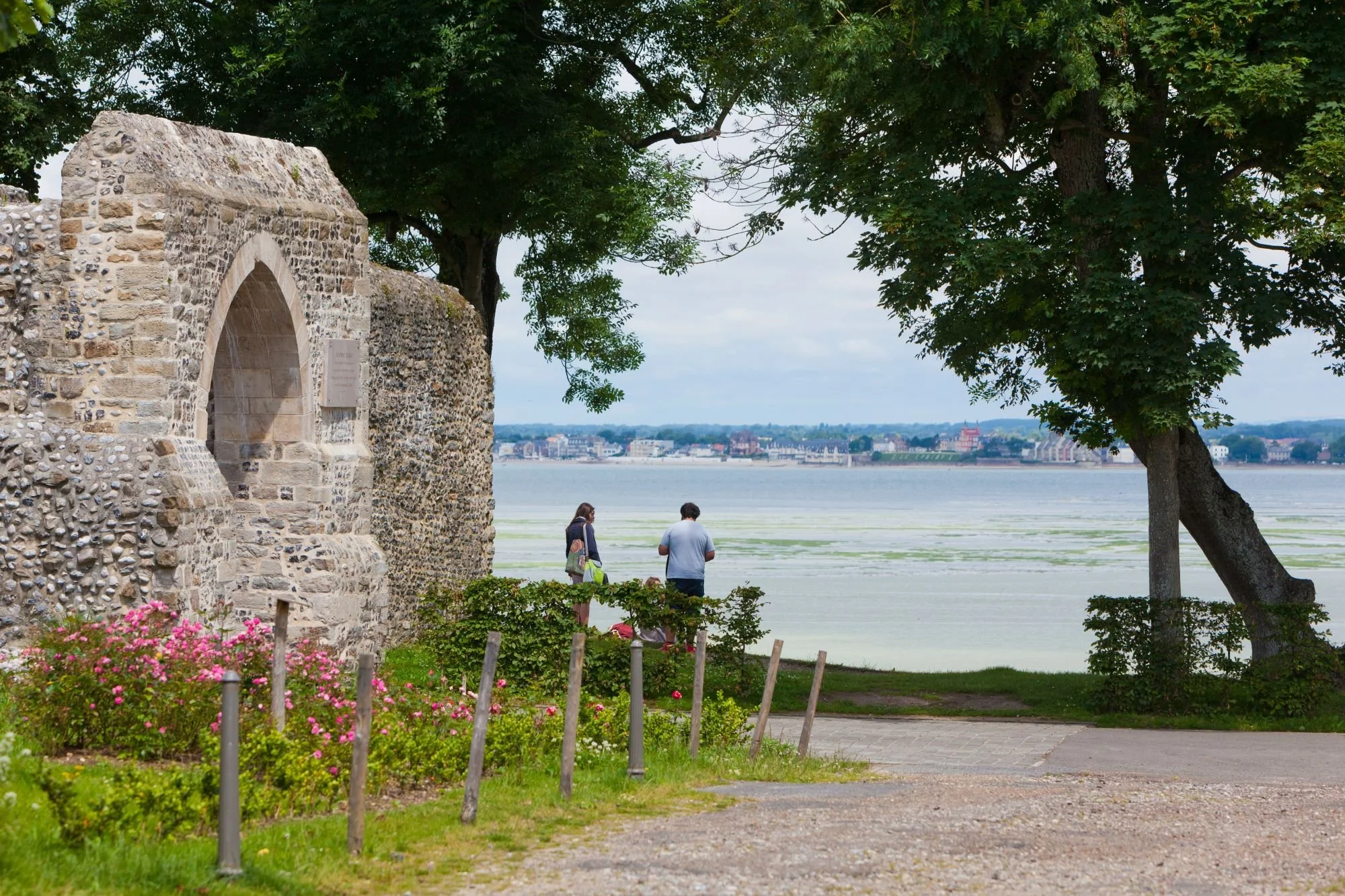 People walking near a historic stone structure by a waterfront with trees and a cityscape in the background.
