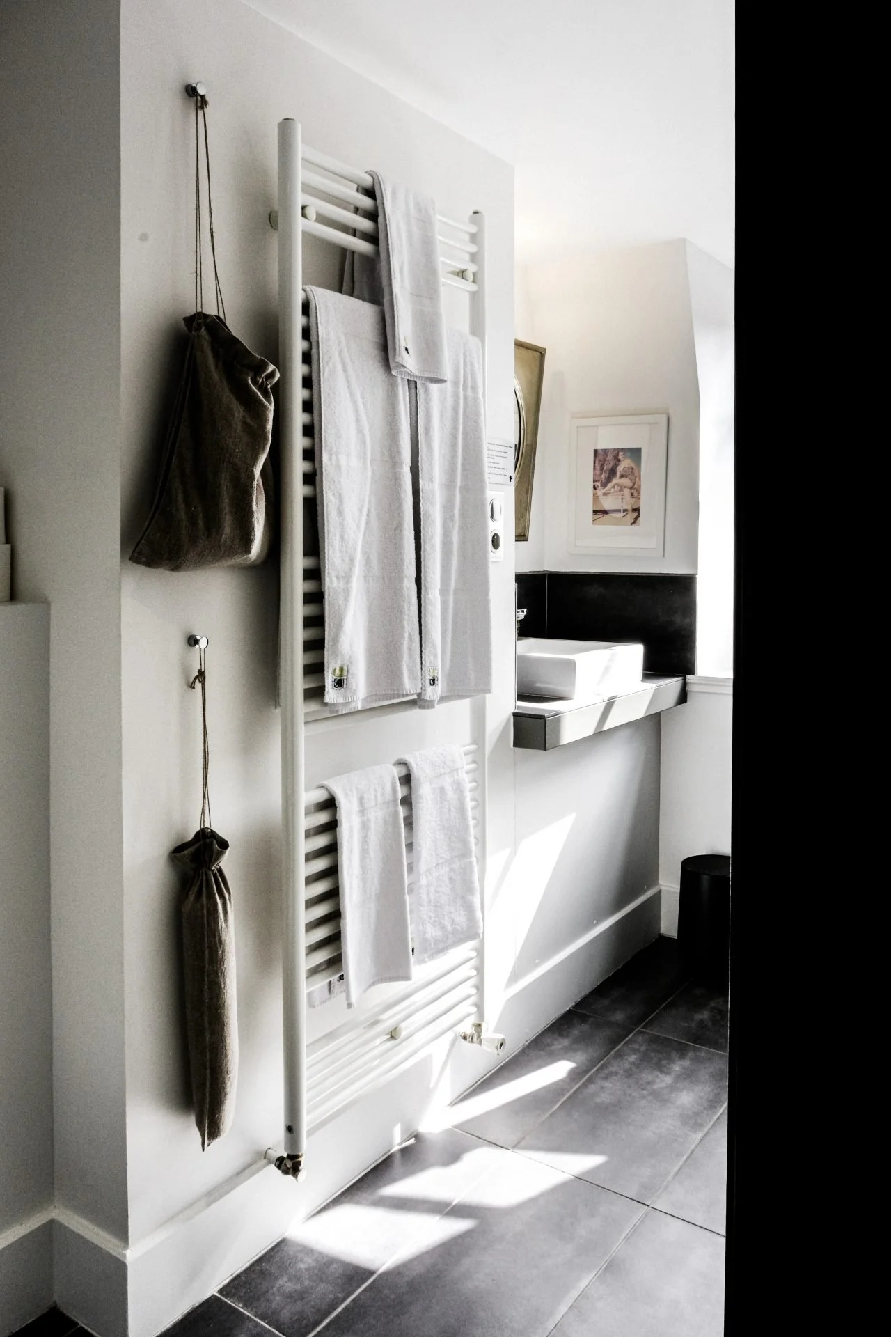 A white towel radiator with white towels hanging on it, a brown bag hanging on the wall, and a sink with a mirror in a bright bathroom with gray tiled floor and white walls.