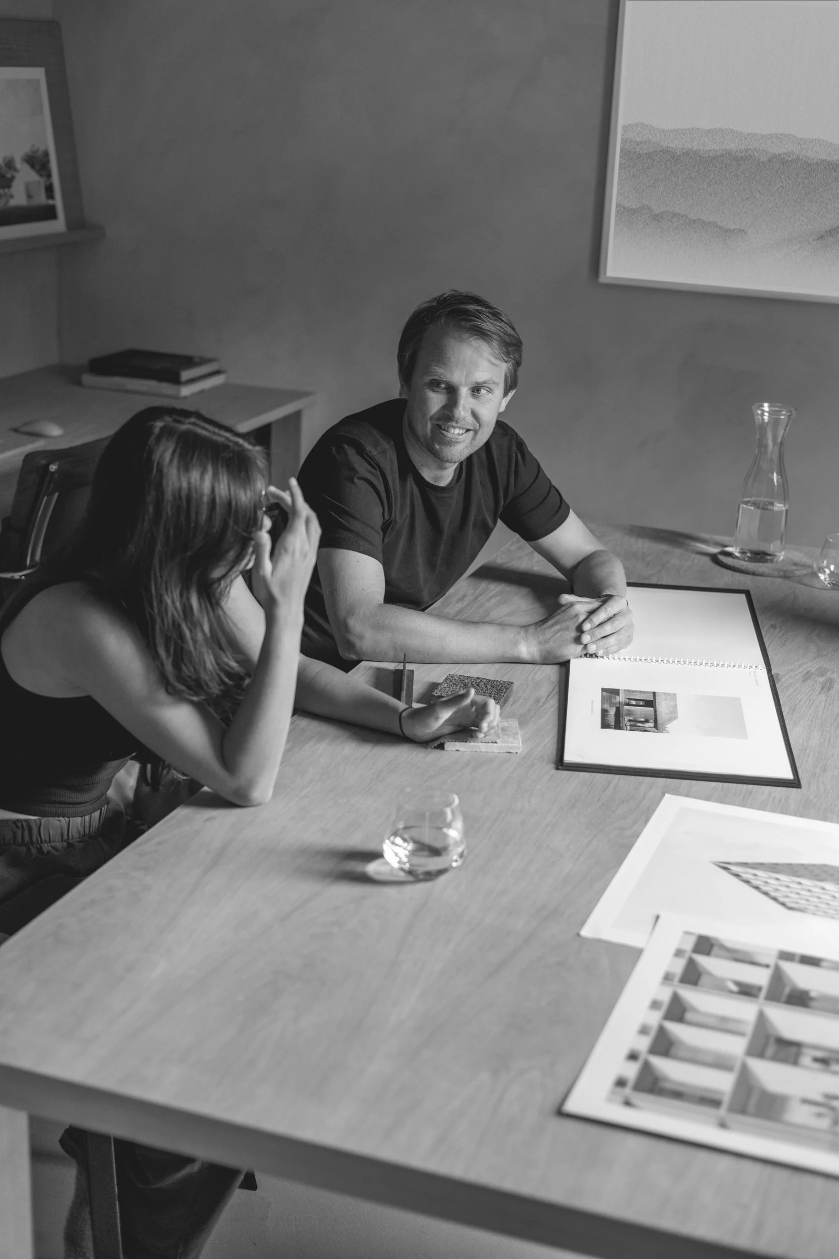 A black and white photo of two people sitting at a wooden table engaged in conversation. The man is smiling and has a sketchbook or notebook in front of him, with a water pitcher and glass nearby. The woman is adjusting her glasses, with photographs and architectural renderings on the table.