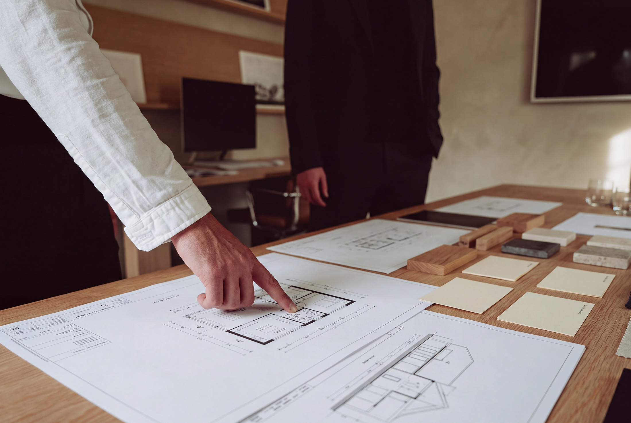 A person pointing at architectural plans on a table with various samples and design documents. Another person in a black suit stands nearby in an office or meeting room with wooden furnishings and framed pictures on the wall.