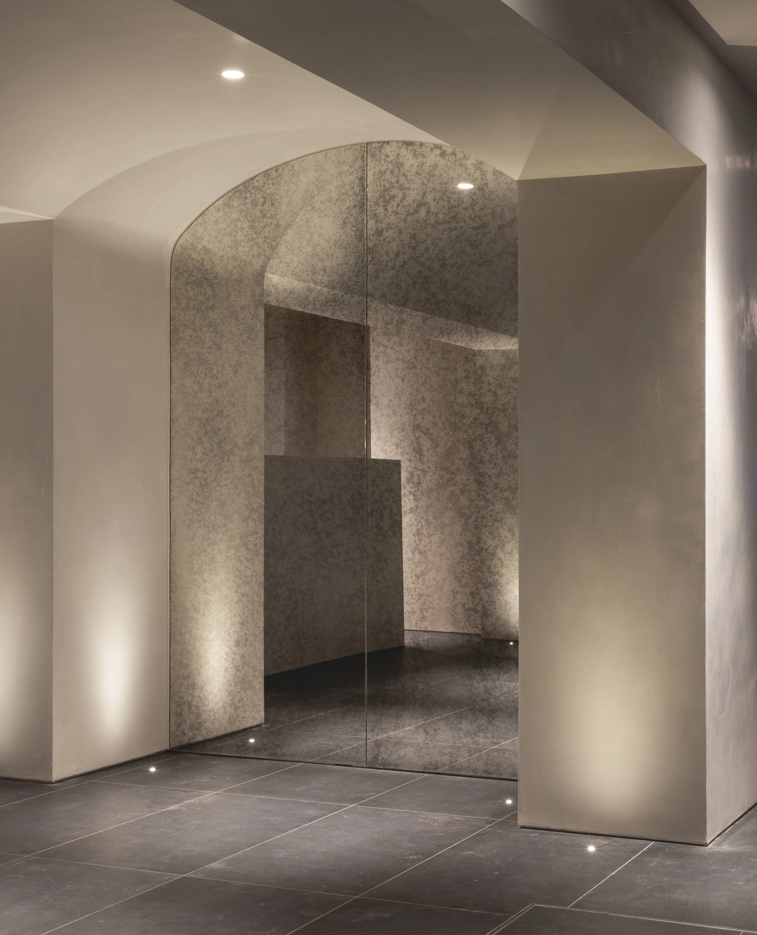 Modern interior space with curved white walls, a mirrored panel, and dark tiled flooring, illuminated by small ceiling lights.