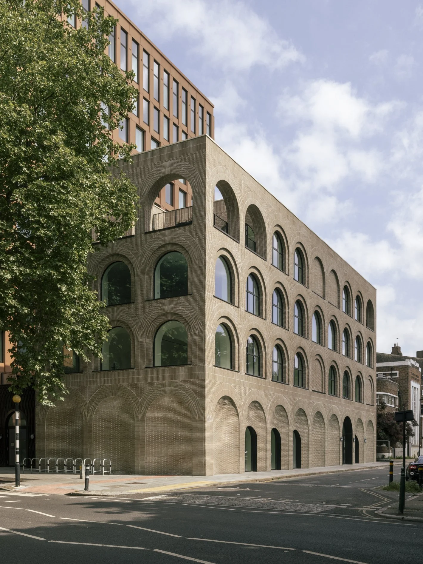 A multi-story building with arched windows and brick facade, partially obscured by a green tree, under a partly cloudy sky, located on a street with marked lanes and traffic barriers.