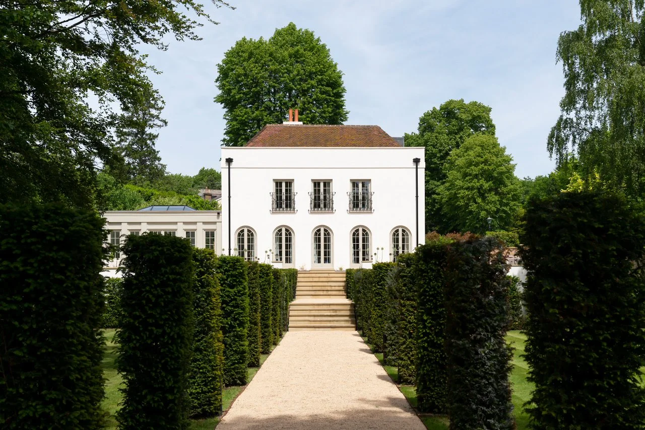 A white two-story house with arched windows and a tiled roof, surrounded by well-kept green trees and shrubs, with a gravel pathway and steps leading up to the entrance.