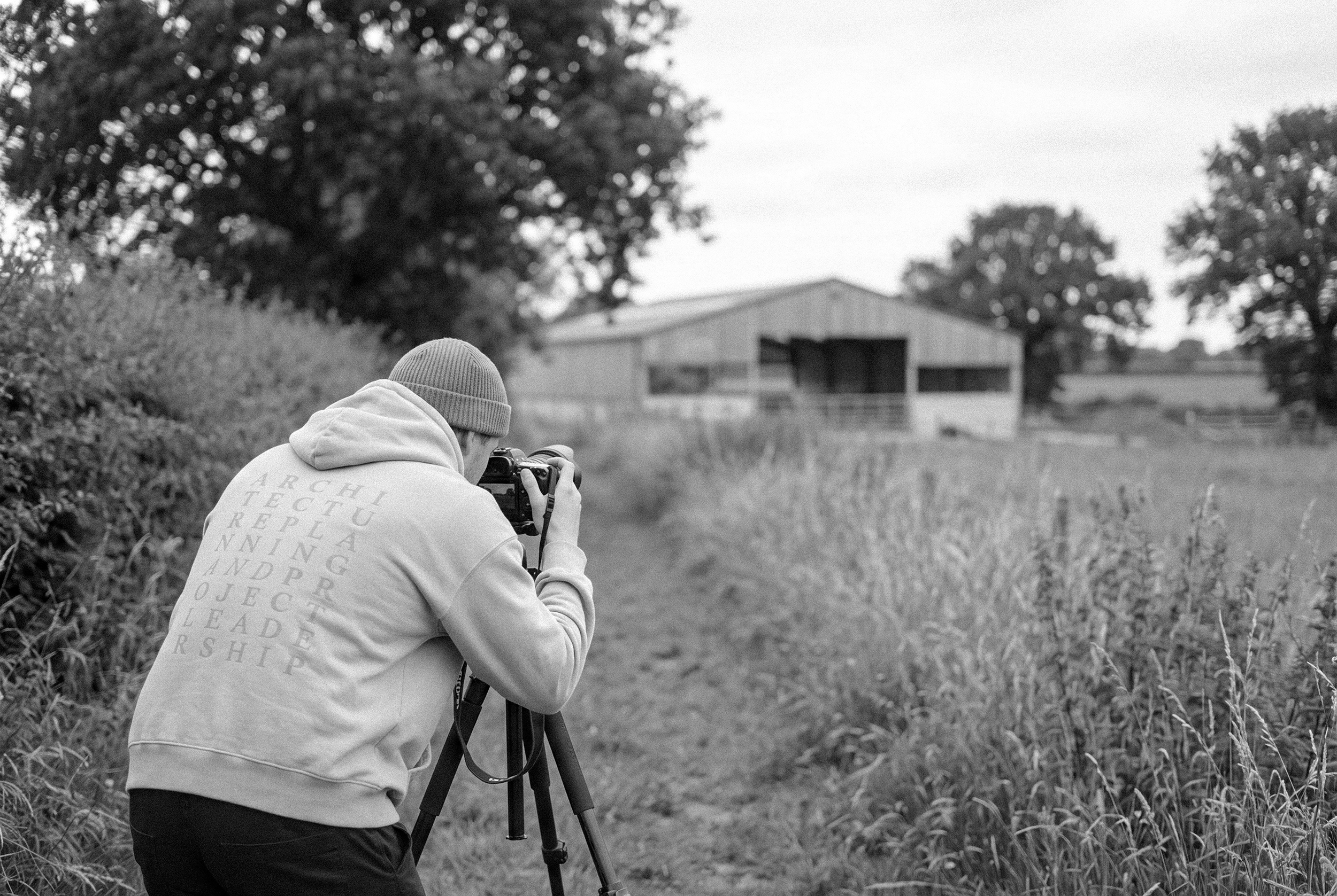 A person wearing a hoodie and beanie is taking a photograph with a camera mounted on a tripod in an outdoor rural setting, with a barn and trees in the background.