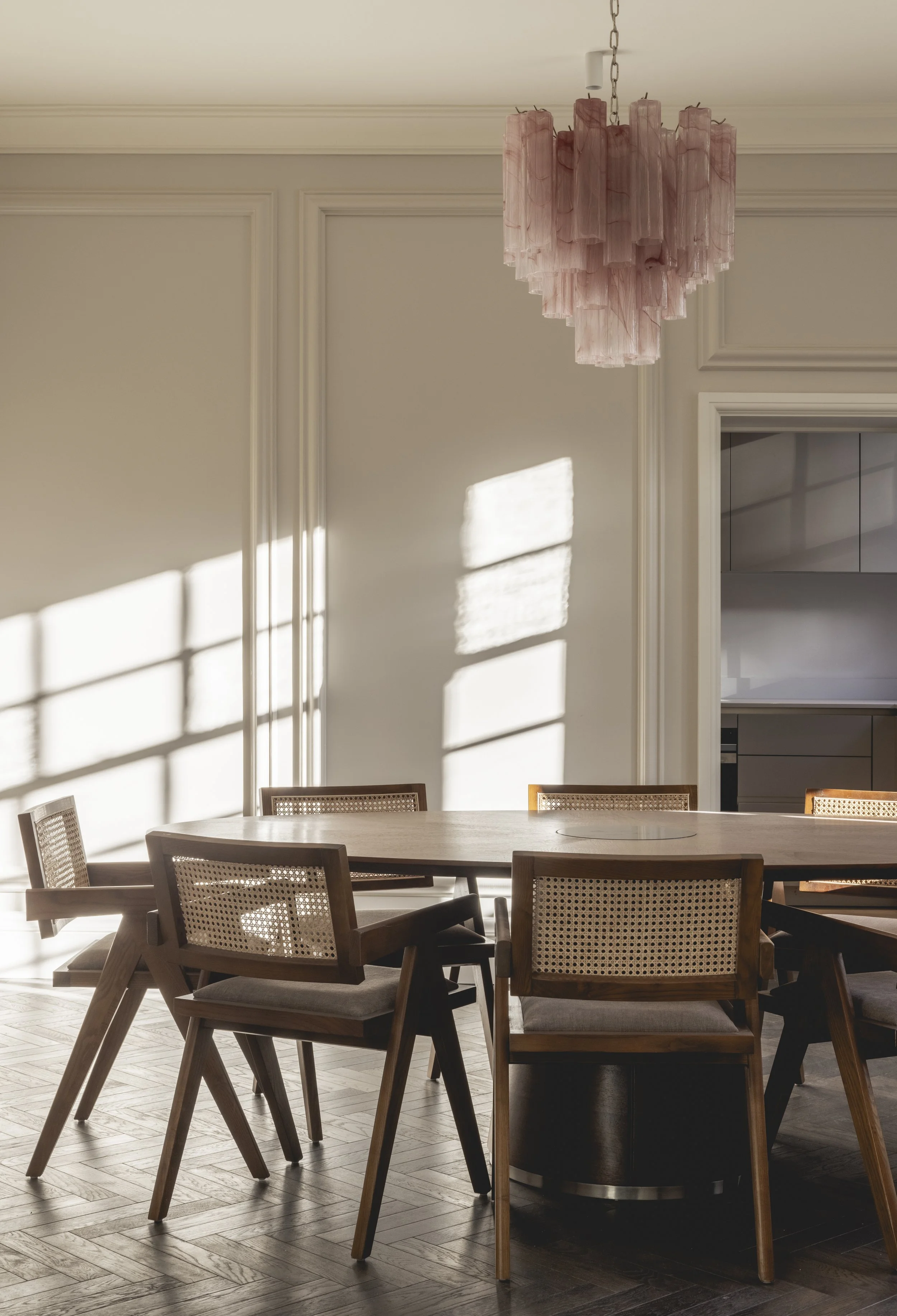 Dining area with a wooden table surrounded by chairs, sunlight casting shadows on the wall, pink chandelier hanging from the ceiling, and partial view of a white cabinet in the background.