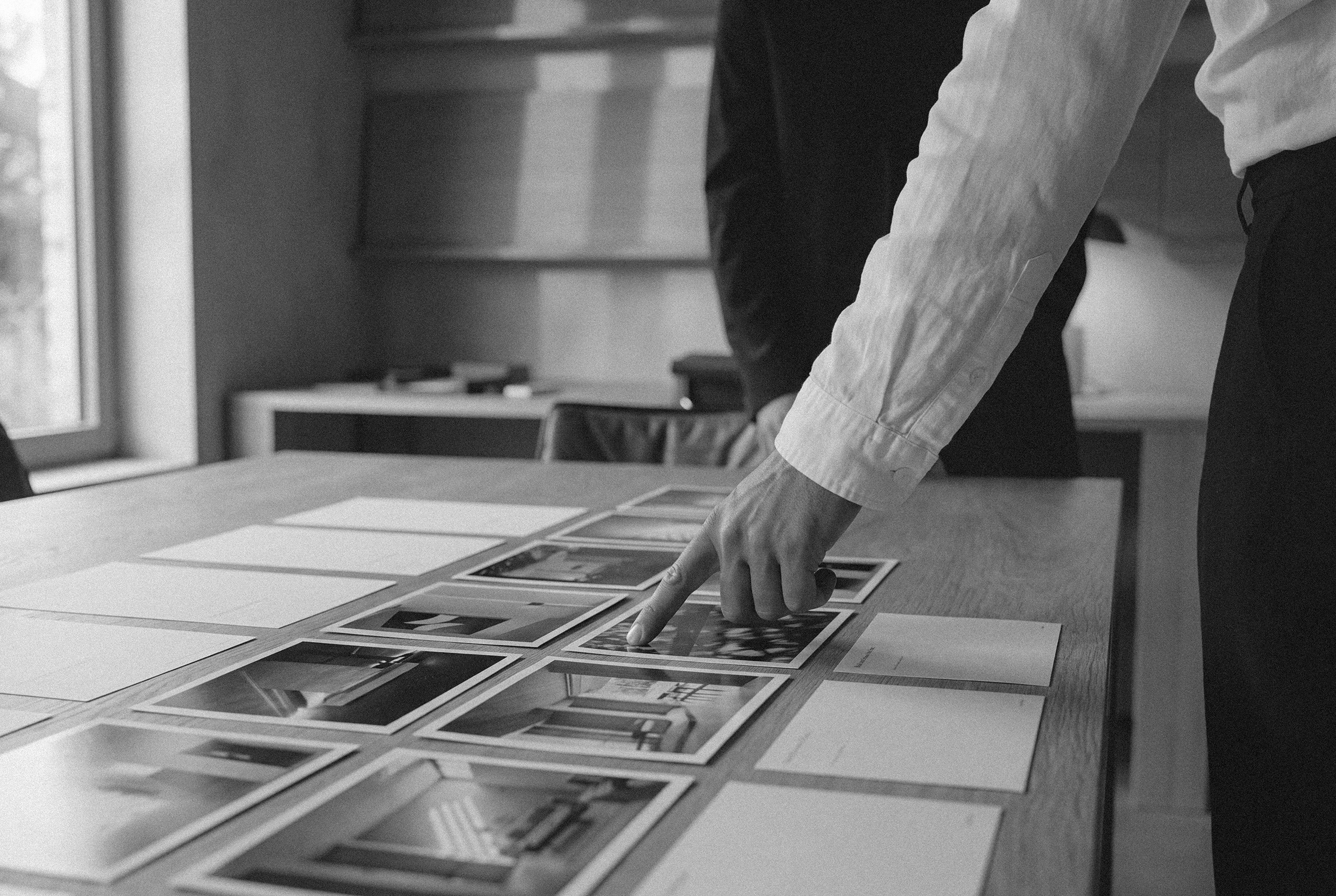 A person pointing at photographs or images laid out on a table during a meeting or discussion, with other people visible in the background.