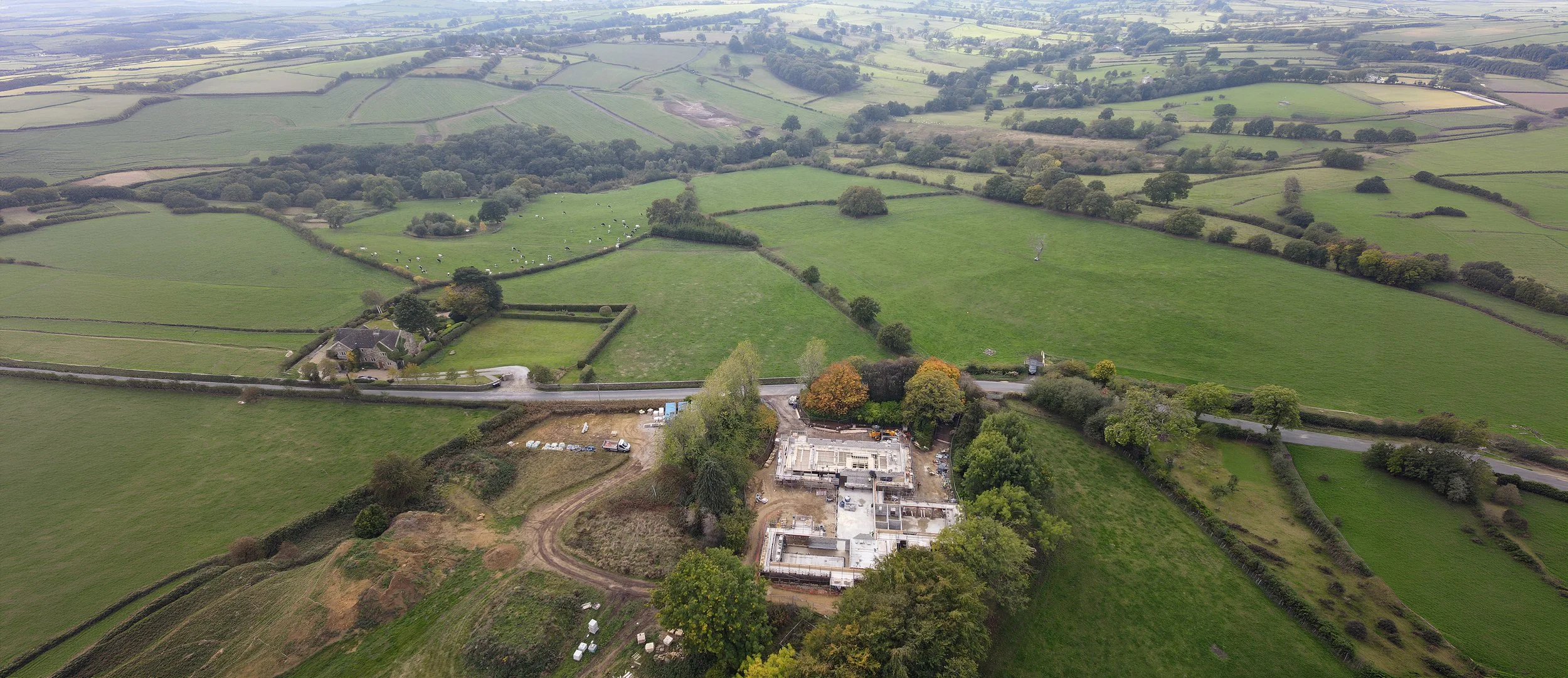 Aerial view of a rural landscape showing green fields, trees, a few houses, and a construction site in the foreground.